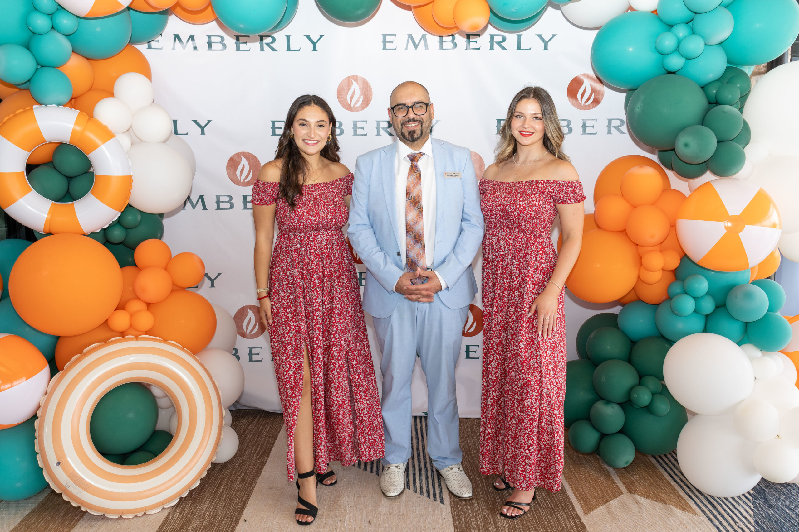 Three people pose in front of an Emberly-branded backdrop at the Grand Opening of Haven Amenity Village, with teal, orange, and white balloon decorations; the man wears a light blue suit, and the women wear matching red dresses.