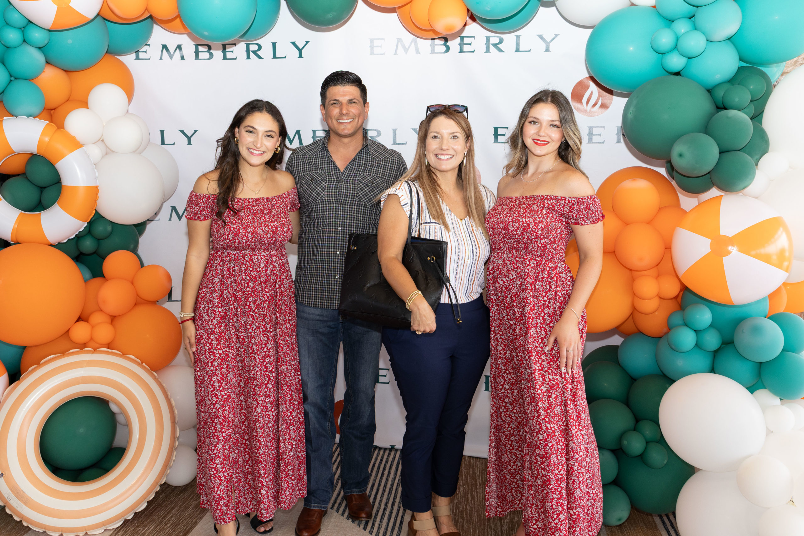 Four people pose in front of a decorated balloon backdrop with “EMBERLY” written on it at the Haven Amenity Village Grand Opening; two women wear matching red floral dresses, surrounded by beach-themed balloons.