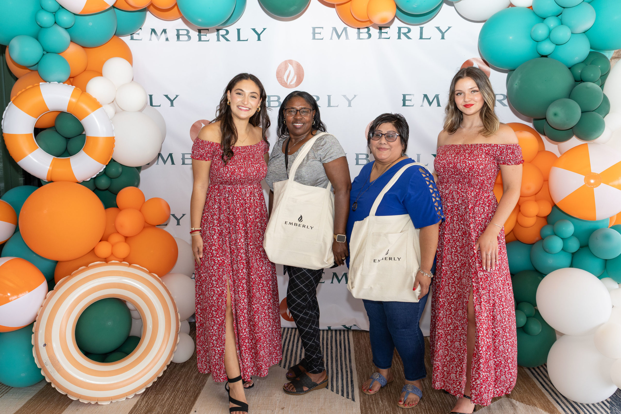 Four women stand in front of an "Emberly" backdrop with colorful balloons and beach balls at the Haven Amenity Village grand opening, two wearing matching red dresses and two holding Emberly tote bags.