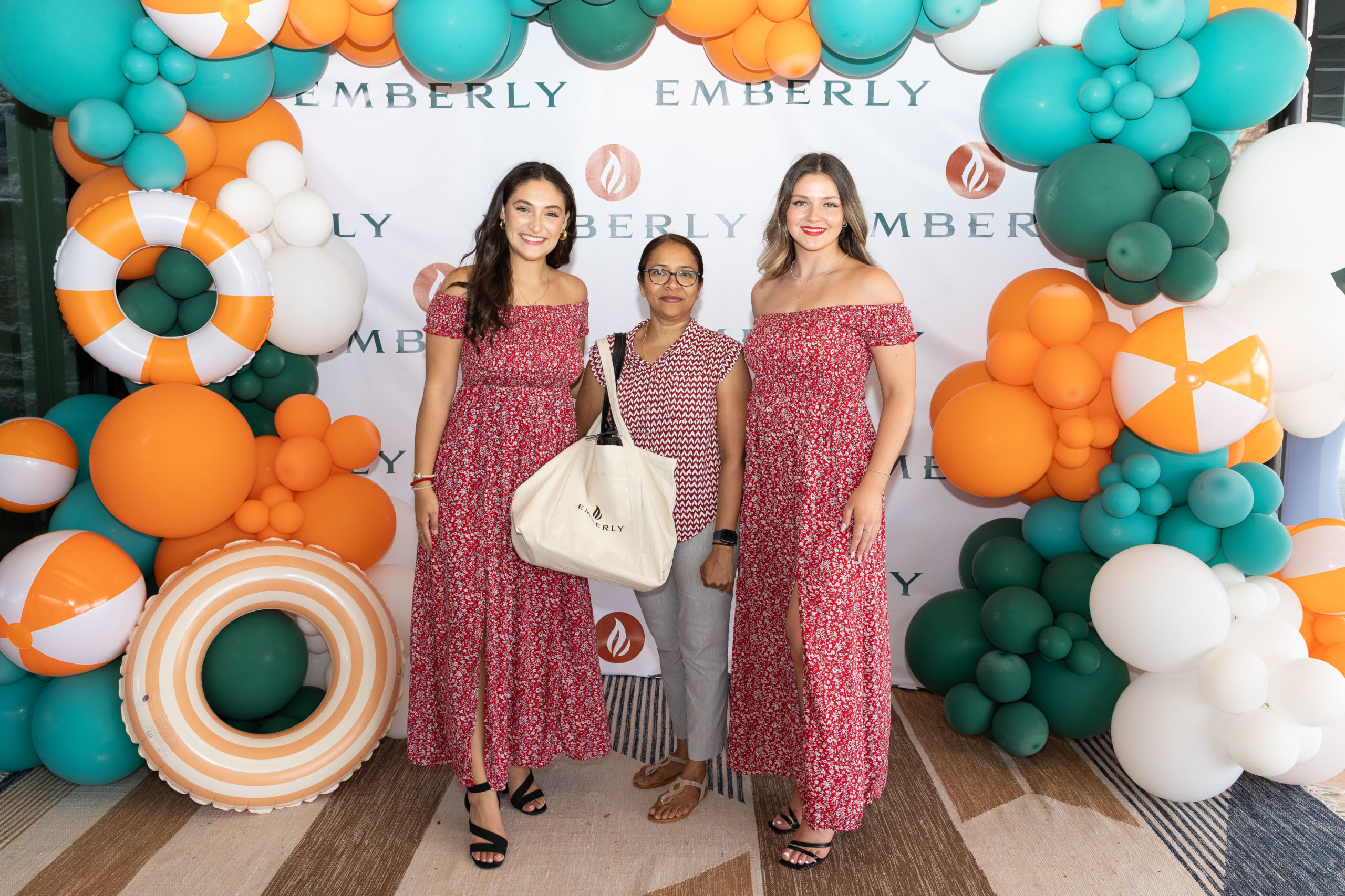 Three women pose in front of an Emberly-branded backdrop at the Haven Amenity Village Grand Opening, decorated with green, orange, and white balloons; two wear matching red dresses, and one holds a tote bag.