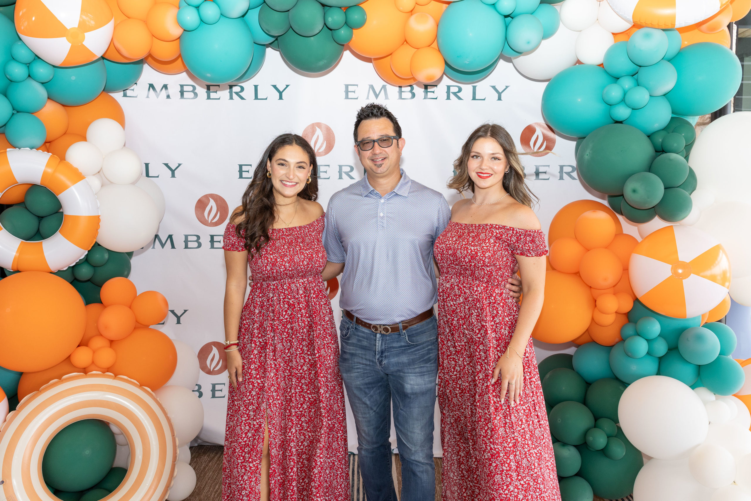 Three people stand in front of a colorful balloon arch with "Emberly" on the backdrop at The Haven Grand Opening. The two women wear matching red dresses; the man wears a blue shirt and jeans.
