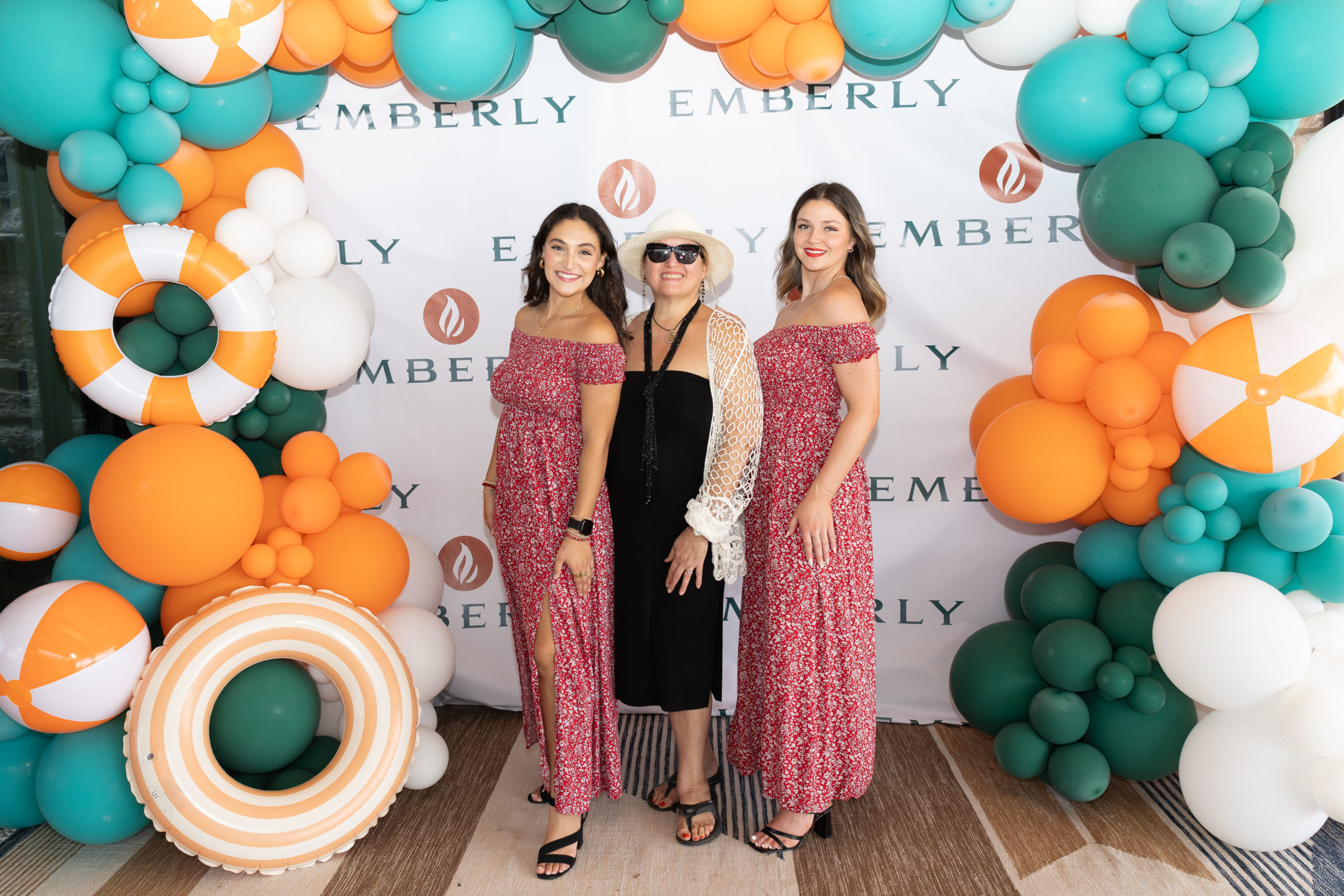 Three women pose in front of an "Emberly" backdrop, decorated with orange, green, and white balloons and beach-themed props, celebrating the Grand Opening at Haven Amenity Village.