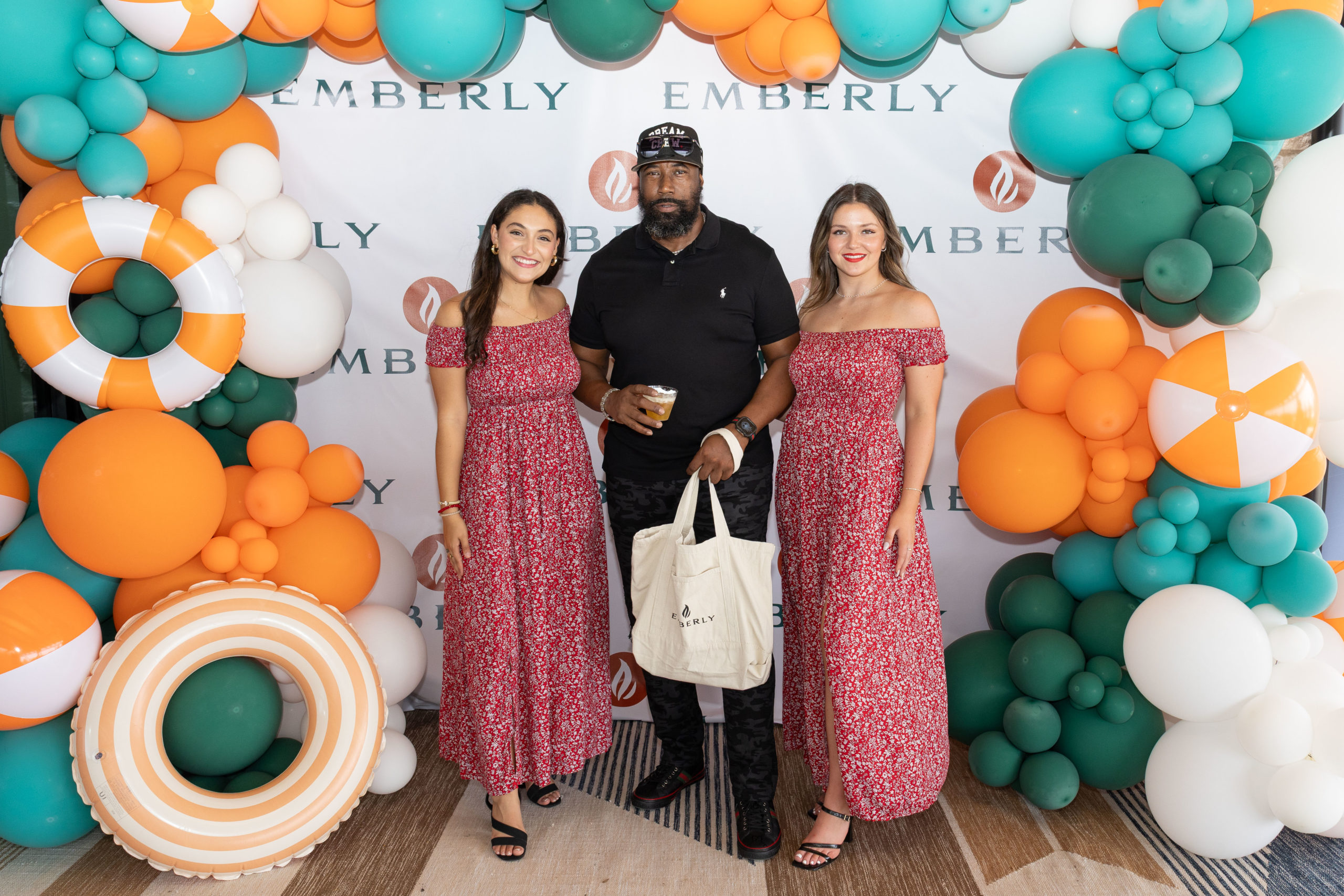 Three people posing in front of an Emberly-branded backdrop at the Grand Opening of Haven Amenity Village, with green, orange, and white balloons; two women in matching red dresses flank a man dressed in black.