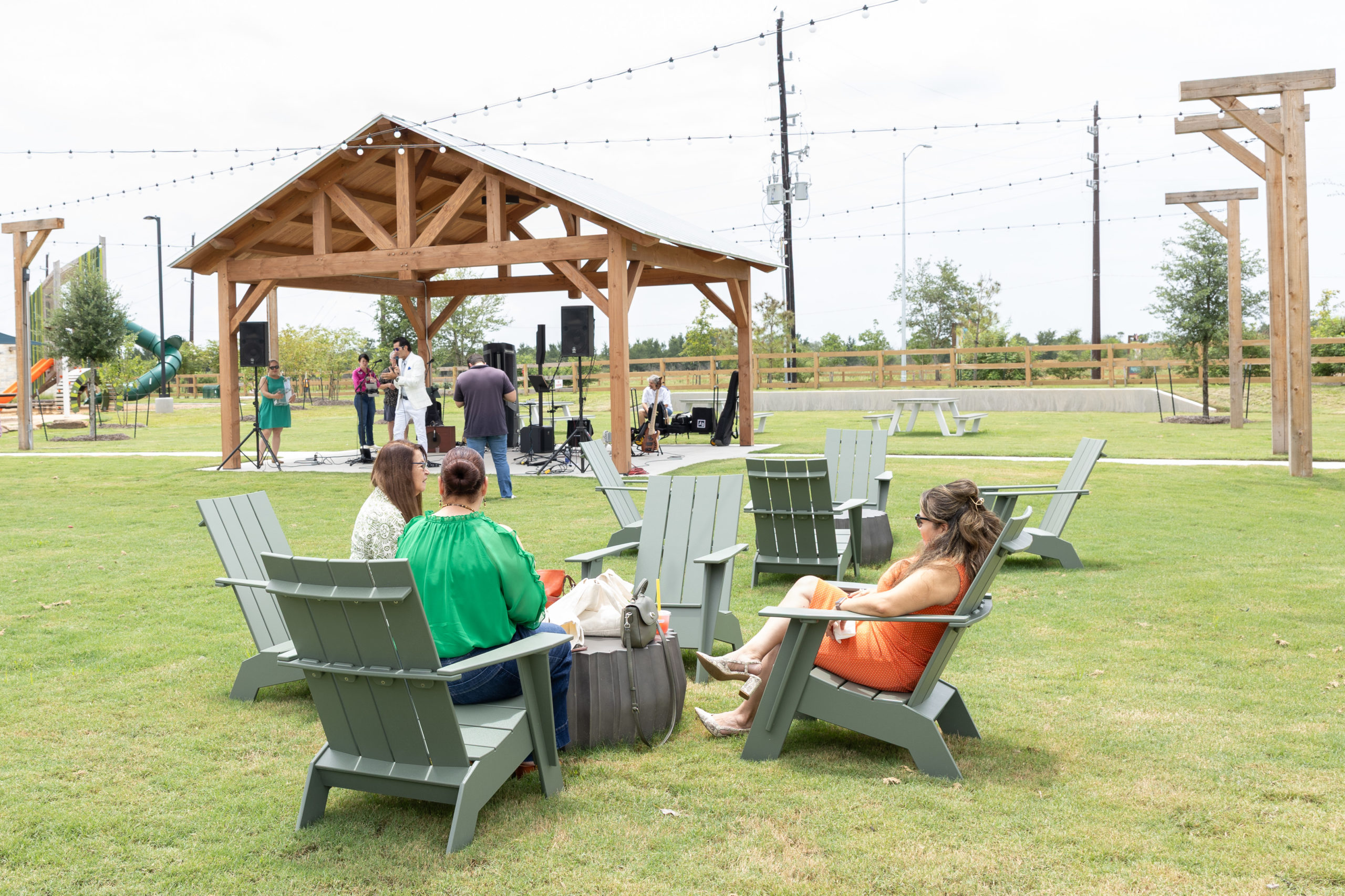 Three women sit on green lawn chairs in a park at the Grand Opening of Haven Amenity Village, watching a small group perform under a wooden pavilion.