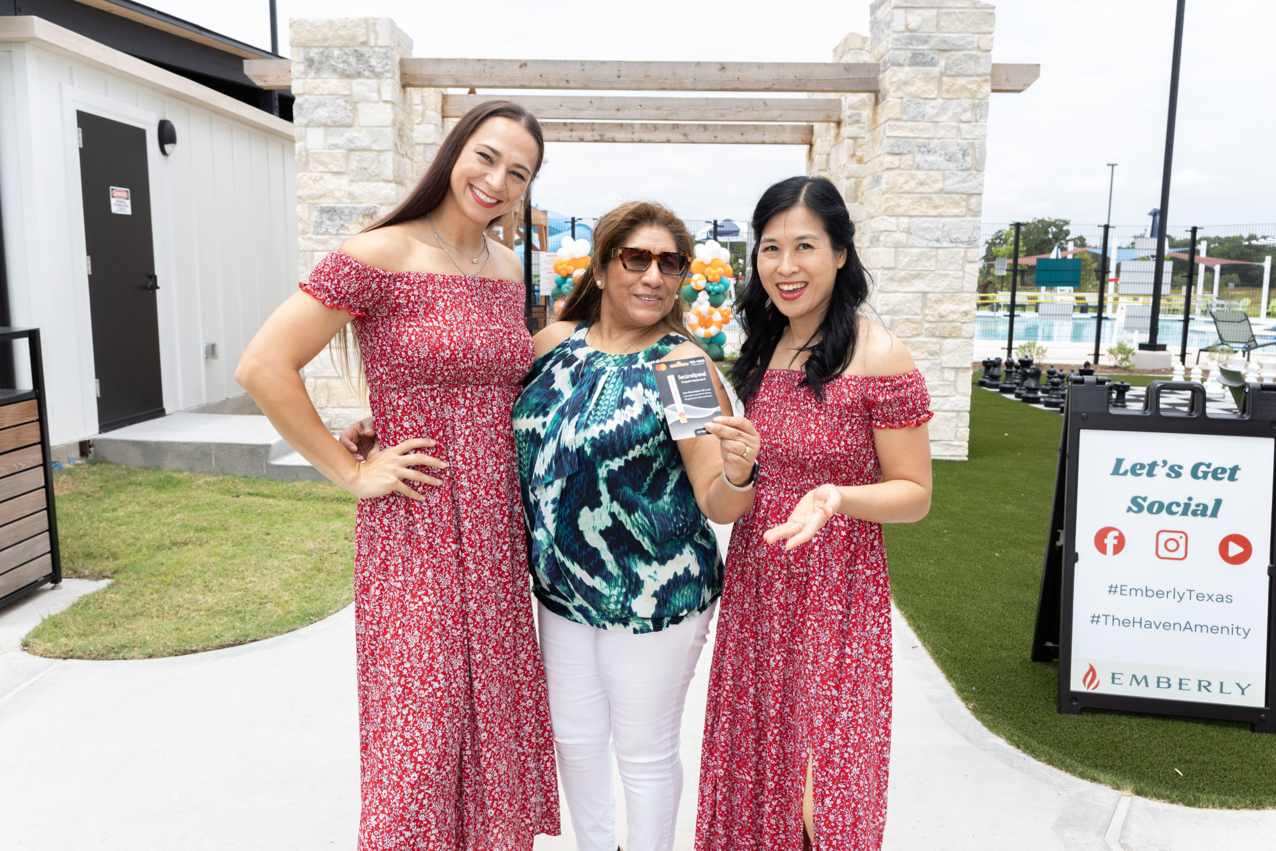 Three women pose outdoors at the Haven Amenity Village Grand Opening; two wear matching red floral dresses, while the center woman, in a blue patterned top, holds a plaque. A sign about social media is visible in the background.