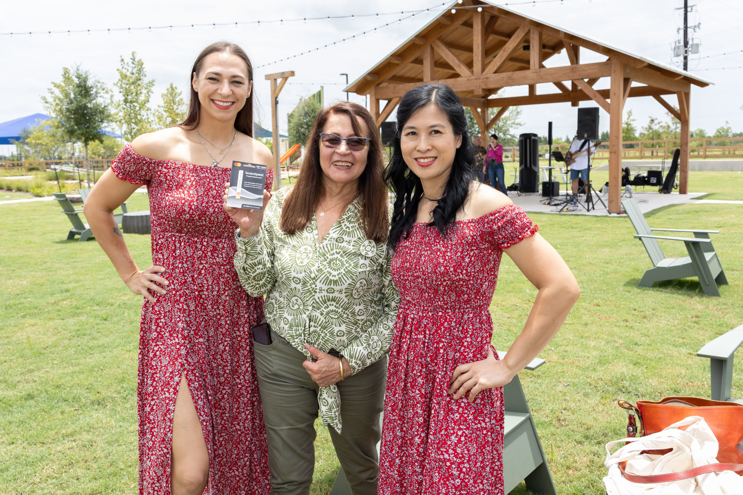 Three women pose outdoors at Haven Amenity Village; two wear matching red dresses, and the woman in the center holds a small item. A wooden pavilion and sound equipment hint at a Grand Opening celebration in the background.