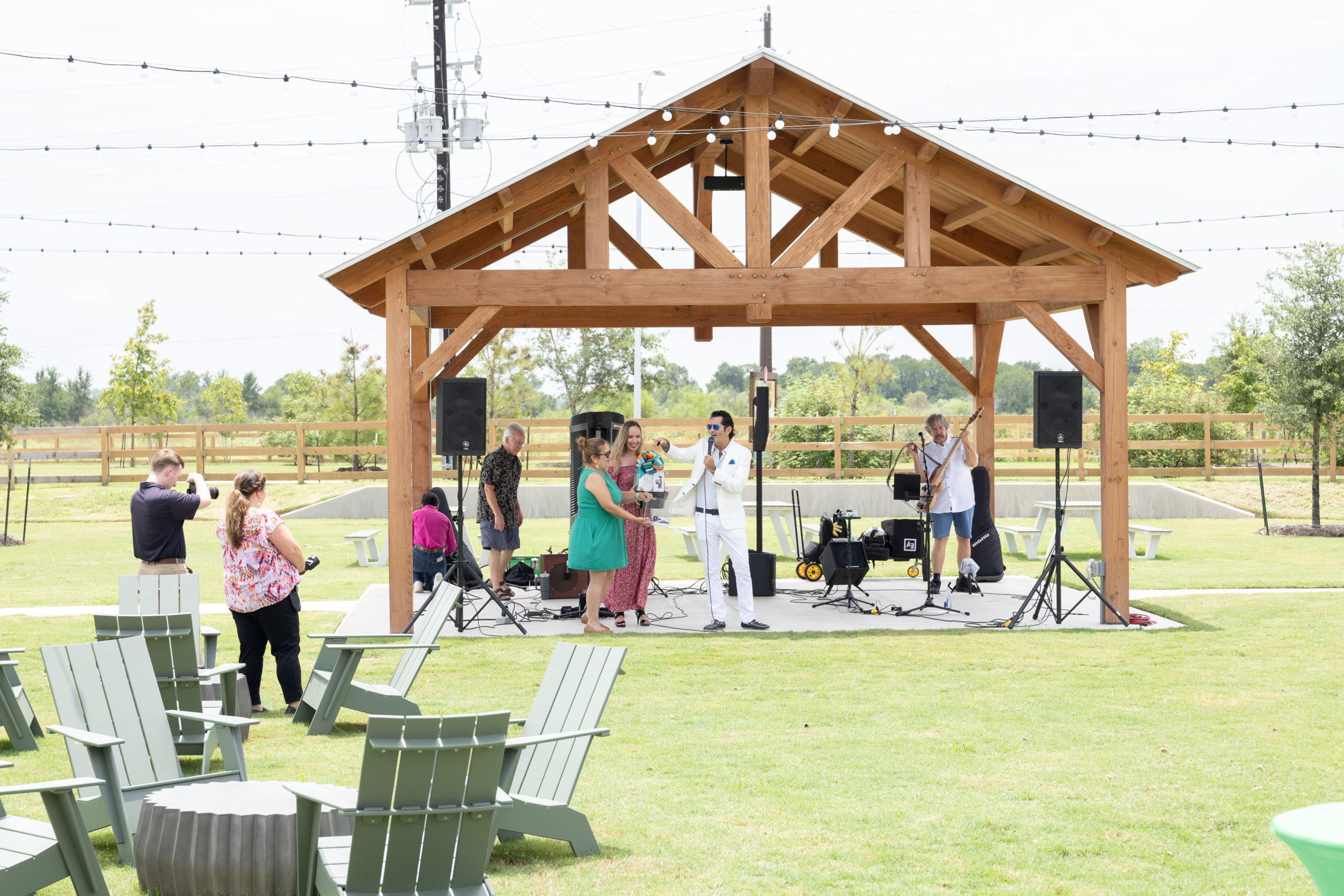 A small band performs on an outdoor wooden stage under a pavilion at the Haven Amenity Village Grand Opening, with a few people watching and taking photos in a grassy area with scattered chairs.