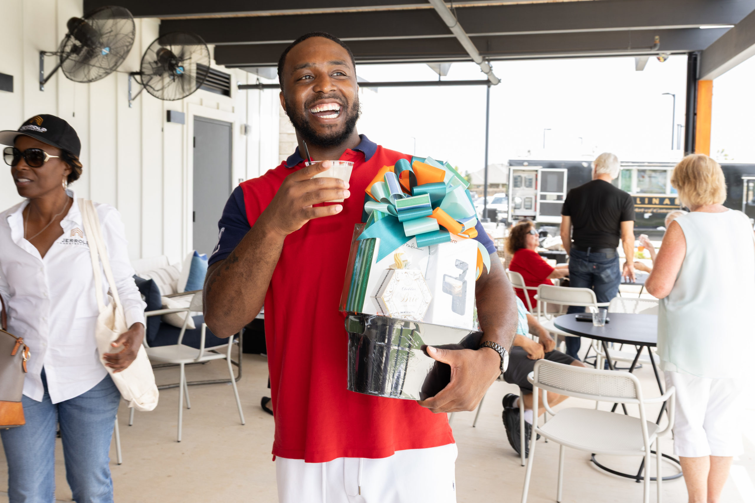 A man in a red shirt smiles while holding a colorful gift basket and a drink at the Haven Amenity Village Grand Opening, with people enjoying the outdoor social event in the background.
