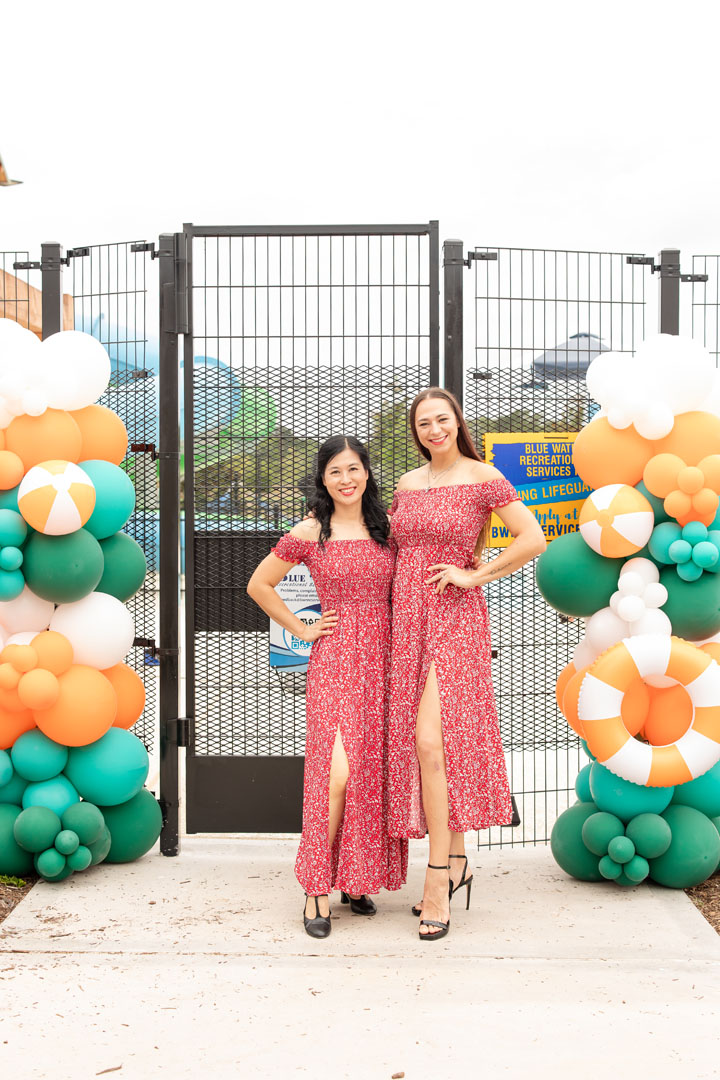 Two women in matching red floral dresses pose and smile in front of a metal gate decorated with green, orange, and white balloons at the Grand Opening of Haven Amenity Village.