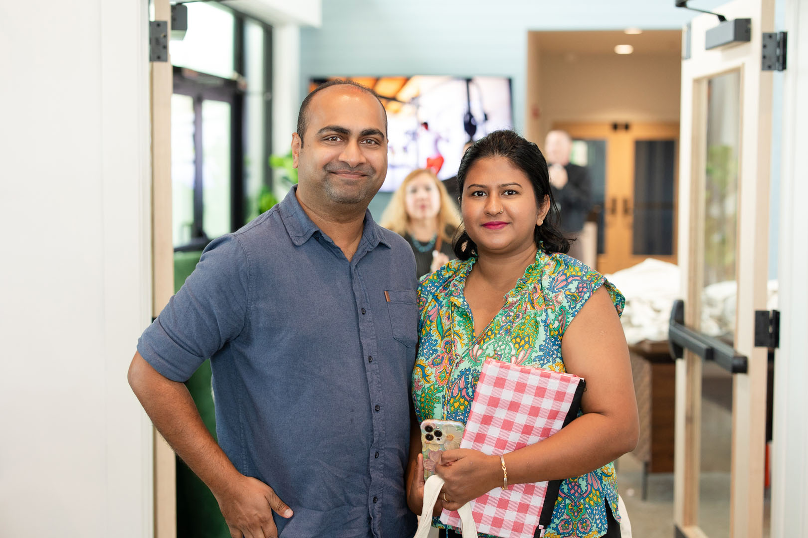A man and a woman stand indoors at the Haven Amenity Village Grand Opening; the man is smiling with his hand on his hip, while the woman holds a phone and a pink-checkered folder, both facing the camera.