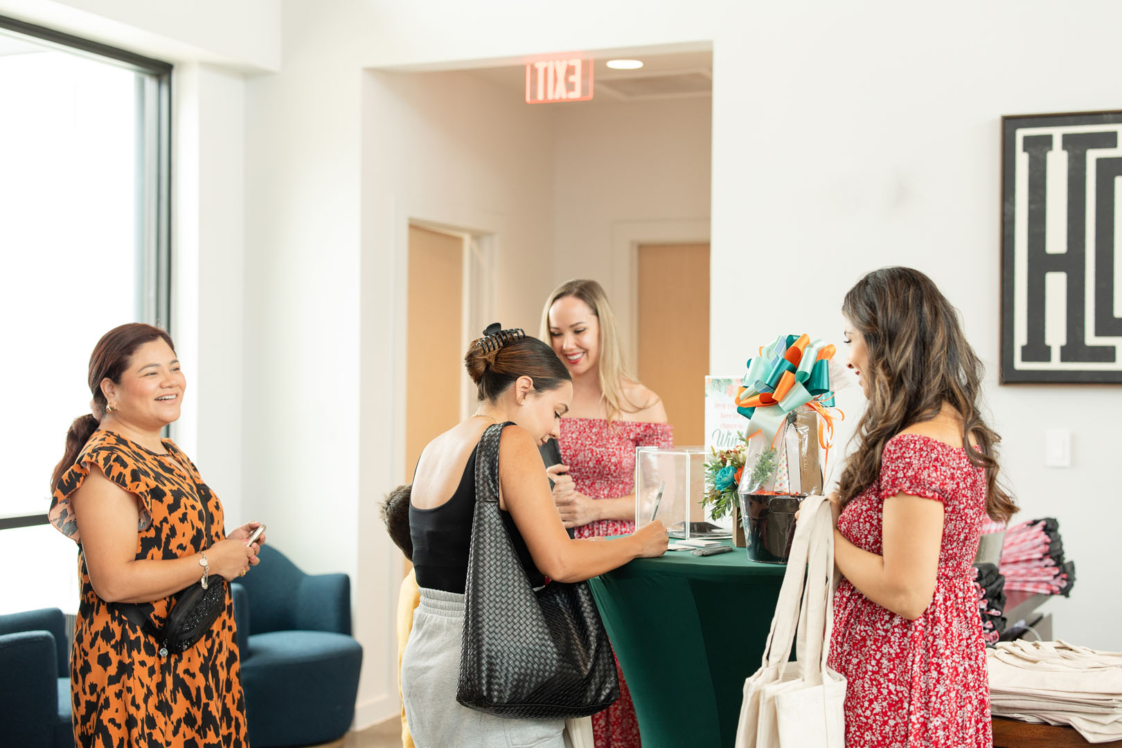 Four women stand around a green table in a bright room at the Haven Amenity Village Grand Opening, chatting and looking at items on the table. One woman smiles while holding a black purse.