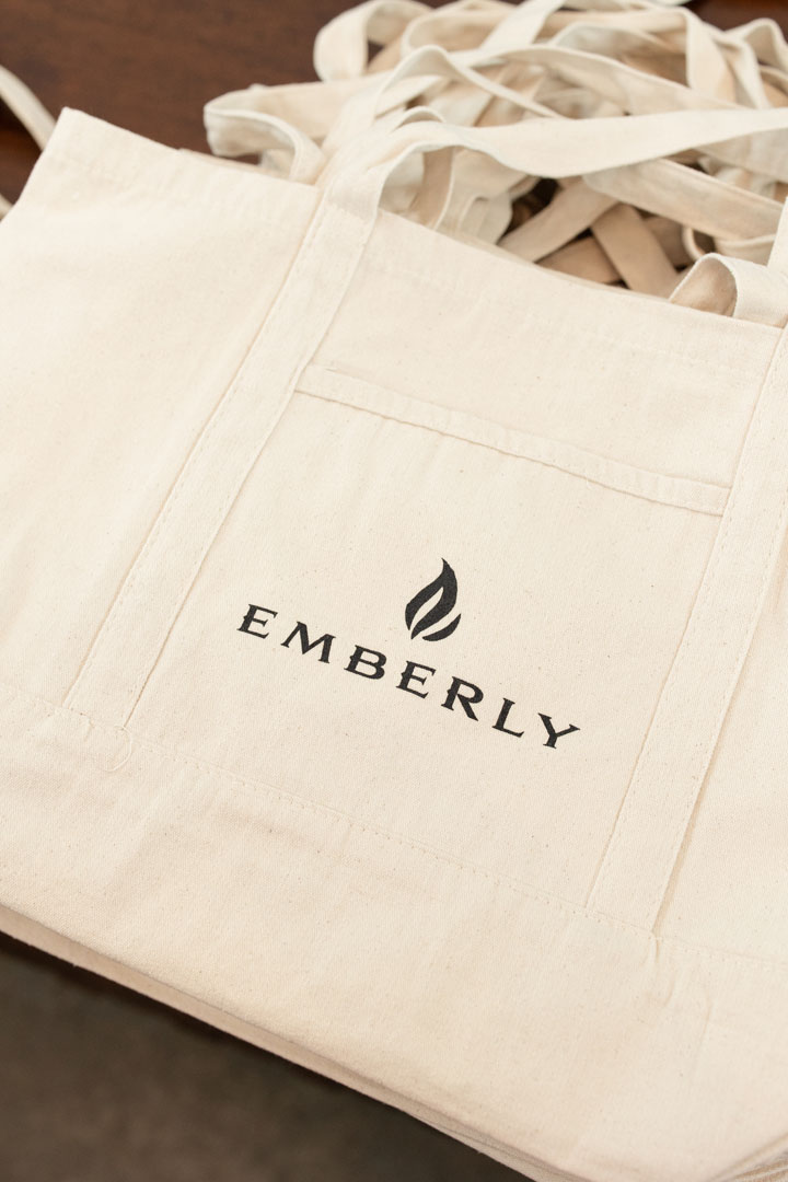 A cream-colored canvas tote bag with the word "EMBERLY" and a flame logo printed on the front pocket, lying on a wooden surface—perfect for the Haven Amenity Village Grand Opening.