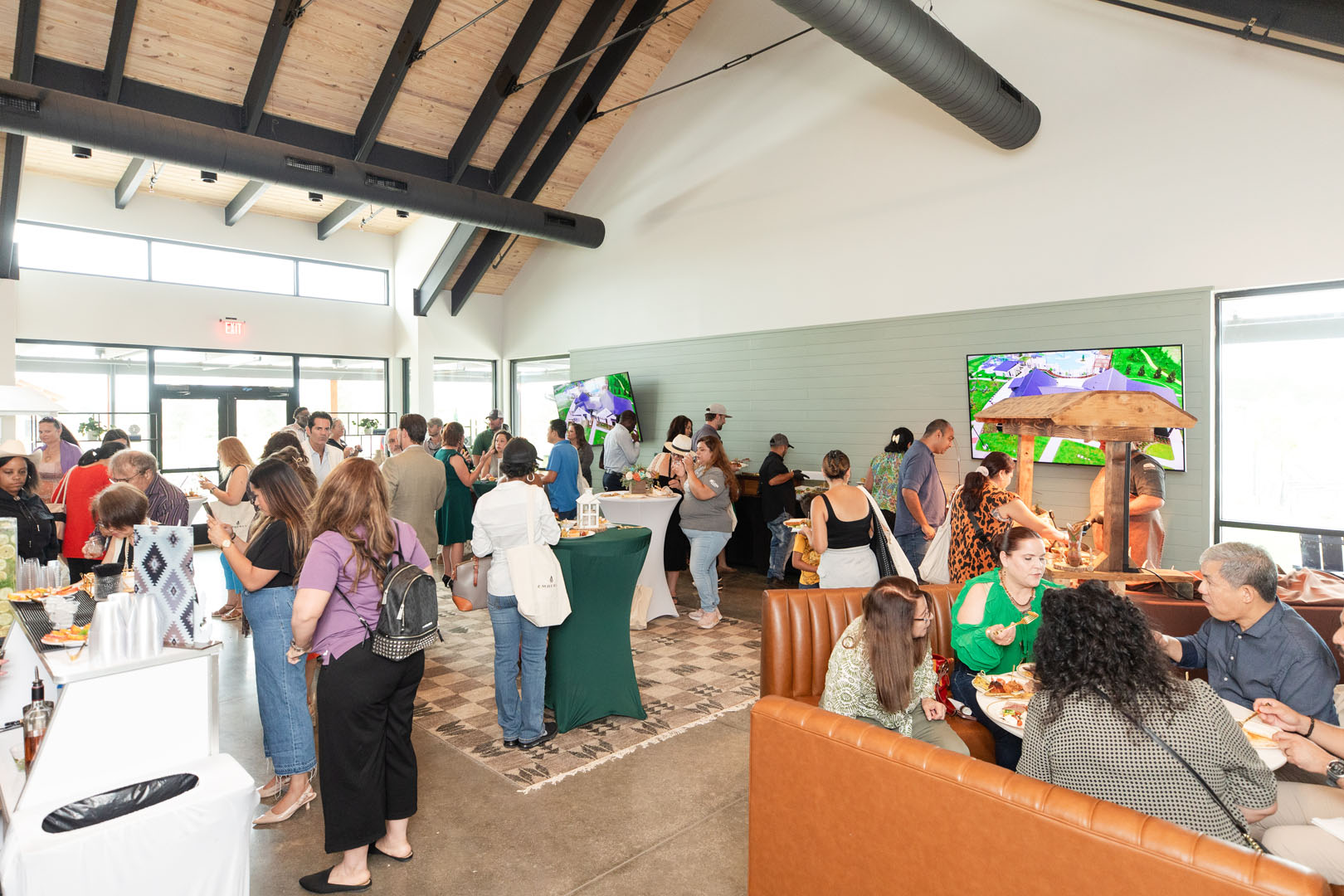 A group of people socializes and eats inside a spacious, modern room with large windows and green-clothed tables during the Haven Amenity Village Grand Opening.