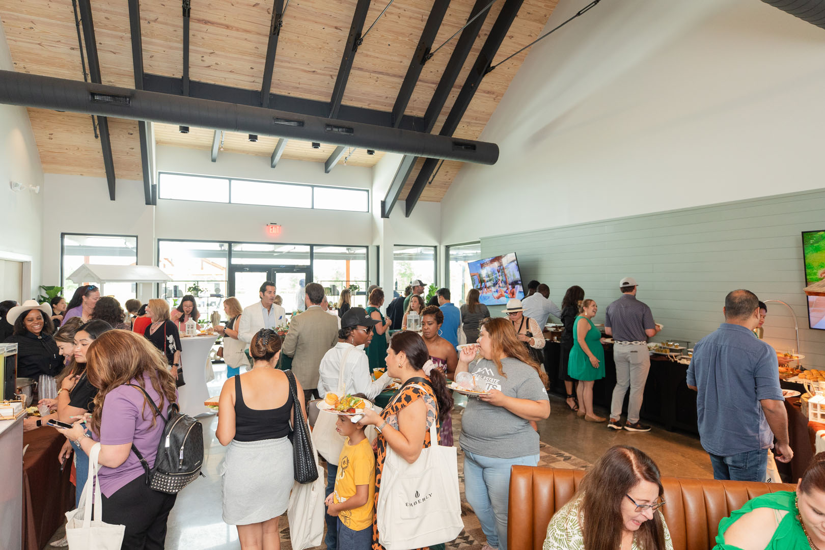 A large group of people socialize and enjoy food at the Grand Opening of Haven Amenity Village, gathered in an indoor event space with buffet tables and high ceilings.