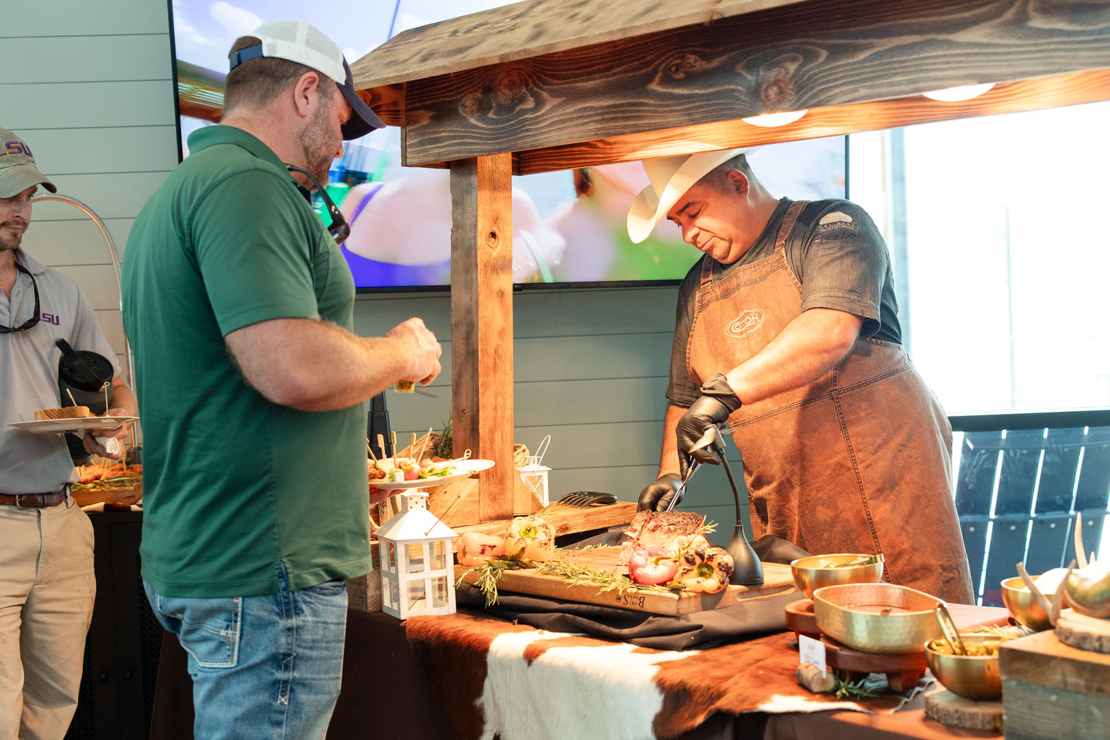 At the Haven Amenity Village Grand Opening, a man in an apron and gloves slices meat at a rustic food station while another man in a green shirt observes. Various food items and serving dishes are displayed on the counter.