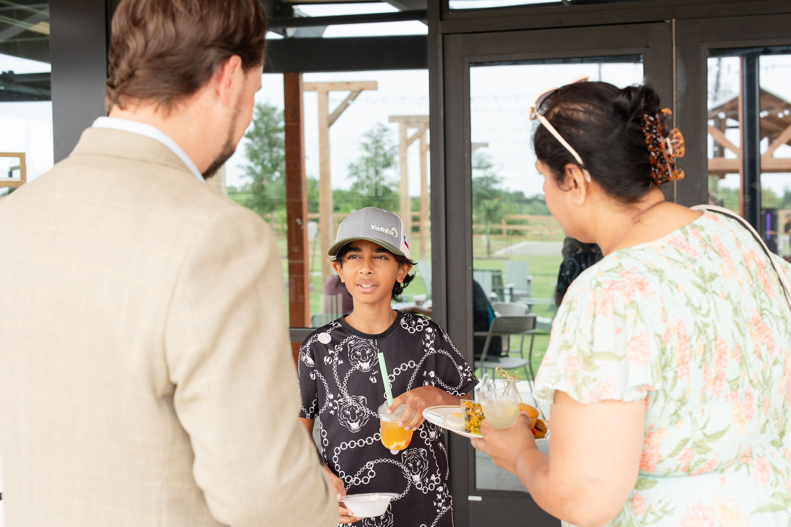 At the Grand Opening of Haven Amenity Village, a boy holding a drink and a plate of food stands between two adults, engaging in conversation outside a building with glass doors.