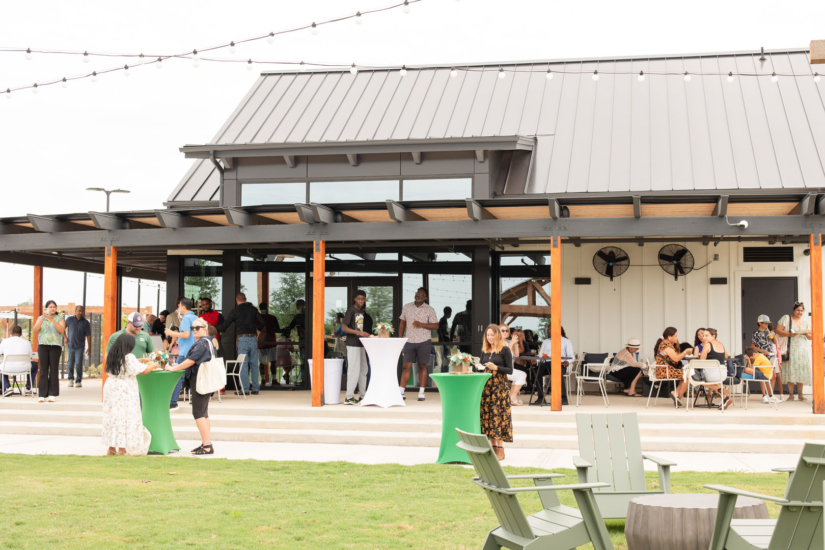 People gather outside Haven Amenity Village’s modern pavilion with string lights overhead, standing and sitting at tables on a lawn during the Grand Opening event.