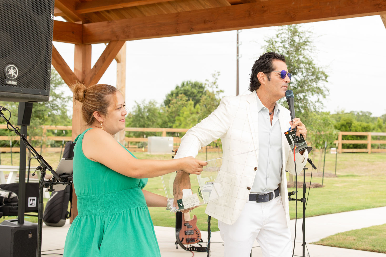 At the Grand Opening of Haven Amenity Village, a man in a white suit speaks into a microphone as a woman in a green dress draws a paper from a clear box at the lively outdoor event.