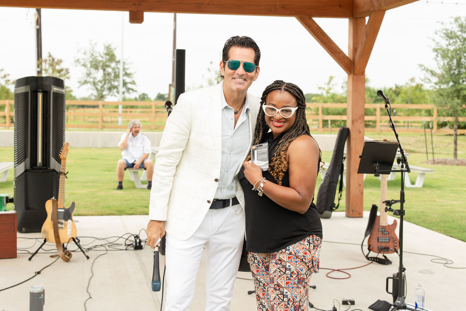 A man in a white suit and sunglasses stands next to a woman holding a badge, both smiling at the Haven Amenity Village Grand Opening, with an outdoor stage and musical instruments in the background.
