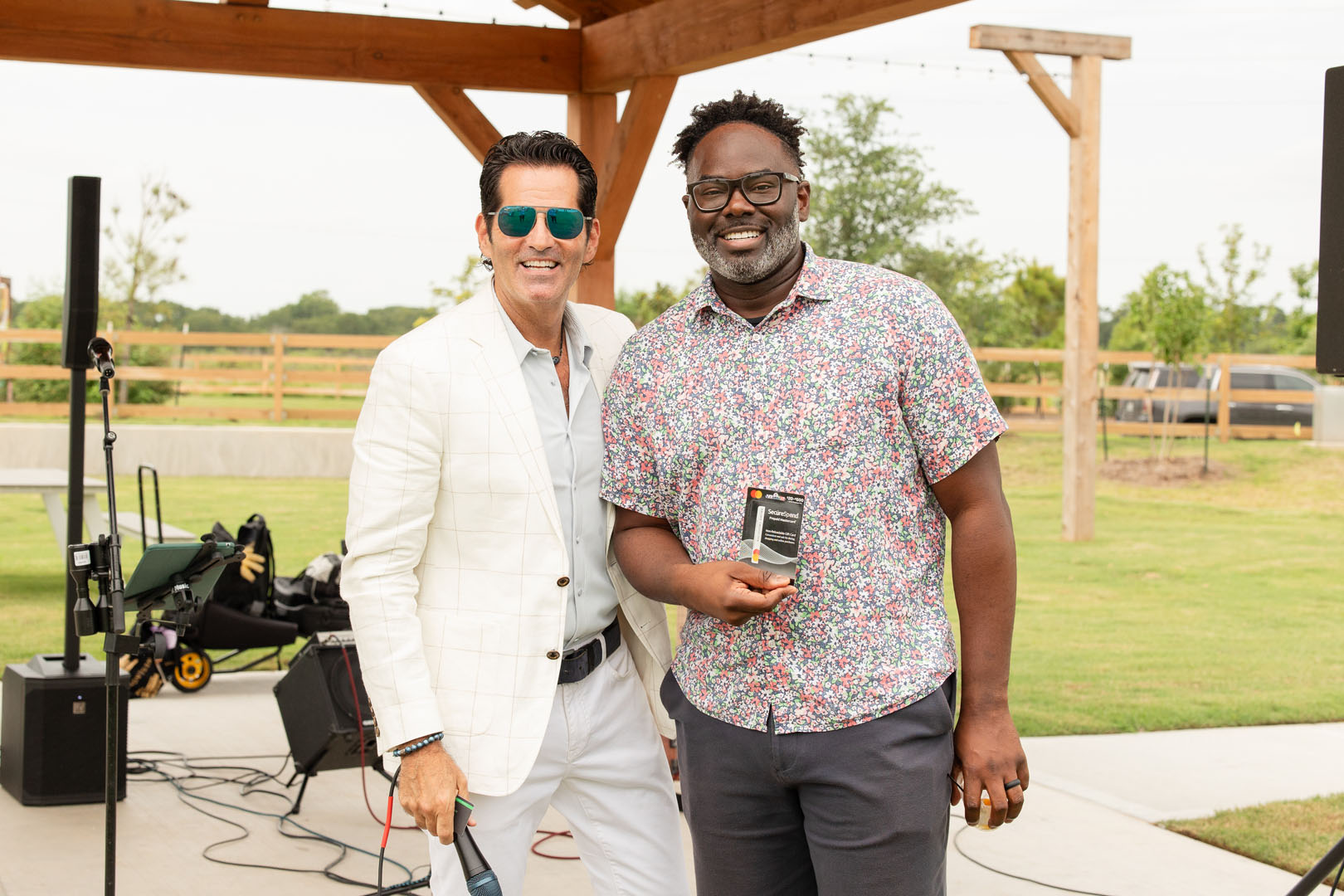 Two men stand side by side outdoors under a wooden pavilion at the Haven Amenity Village Grand Opening; one holds a microphone, the other holds a plaque, both smiling at the camera.