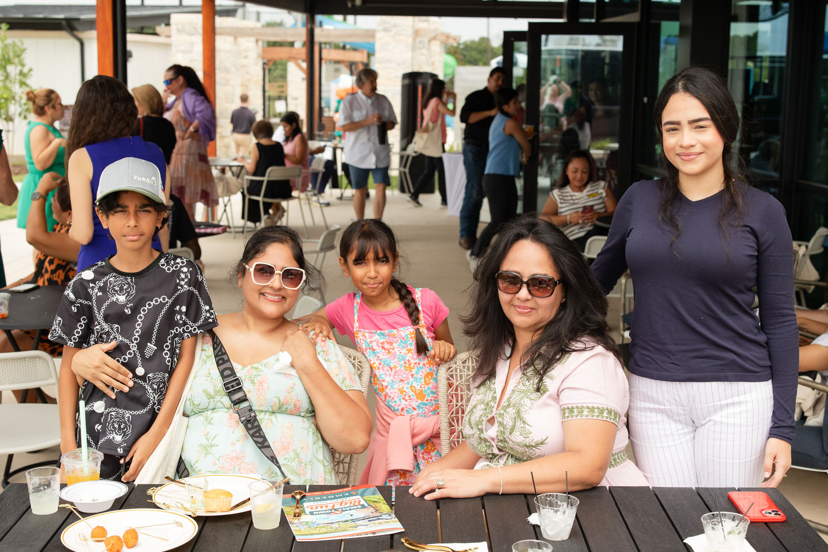 A group of five people, including two children and three adults, pose together at an outdoor restaurant table with drinks and empty plates during the Haven Amenity Village Grand Opening. Other diners are visible in the background.