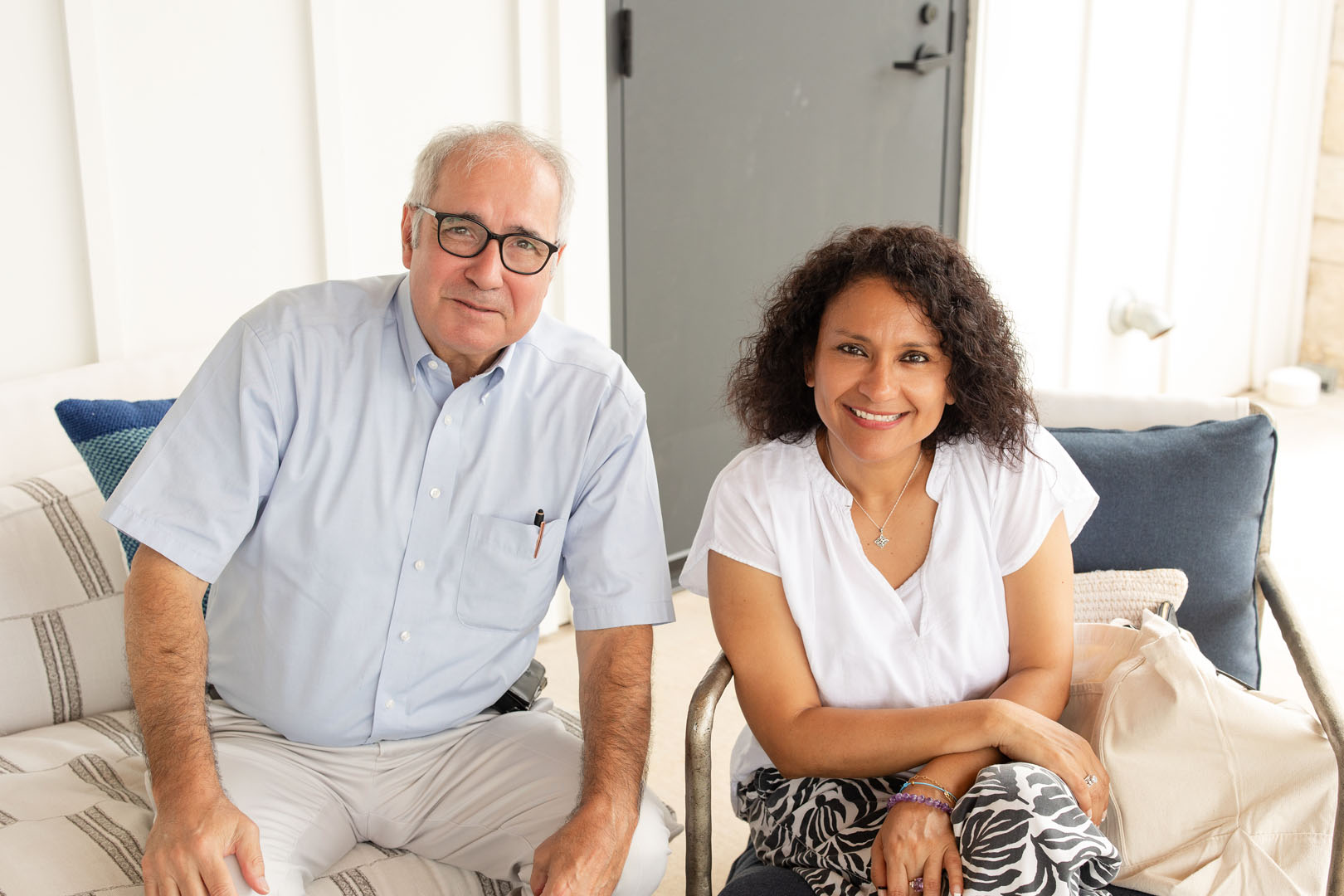 An older man and a woman sit on a sofa indoors at Haven Amenity Village, both smiling at the camera. The man wears glasses and a light blue shirt; the woman has curly hair and a white top.