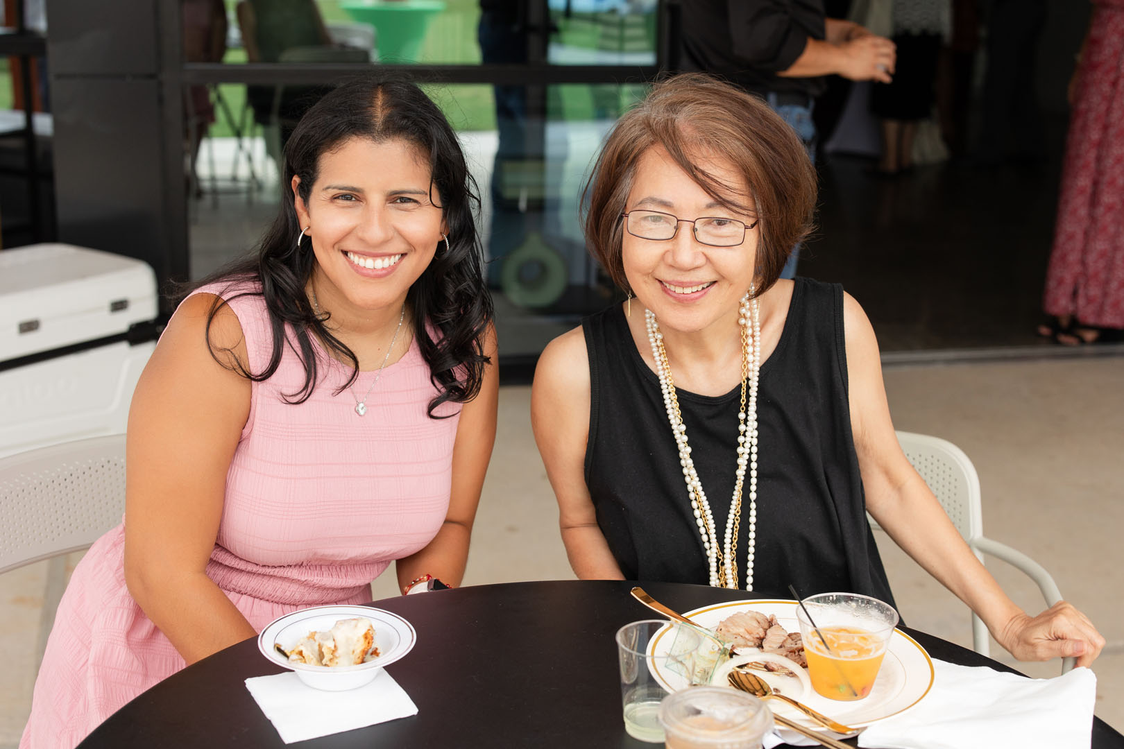 Two women sit at a table with plates of food and drinks, smiling at the camera during the Haven Amenity Village Grand Opening; one wears a pink dress, the other a black top and pearl necklace.