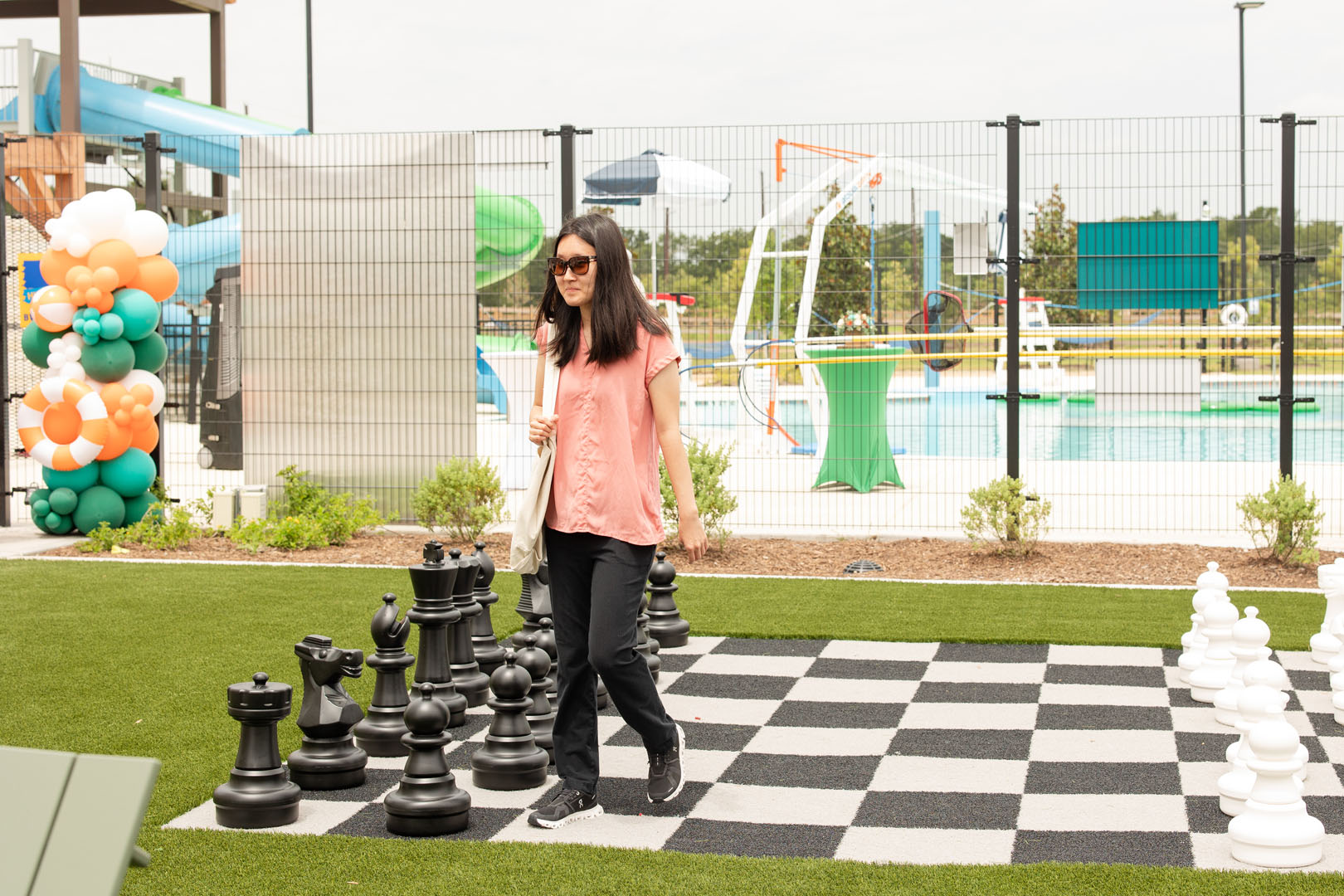 A woman wearing sunglasses walks across a large outdoor chessboard at Haven Amenity Village, near a swimming pool and colorful balloon decorations during the Grand Opening.