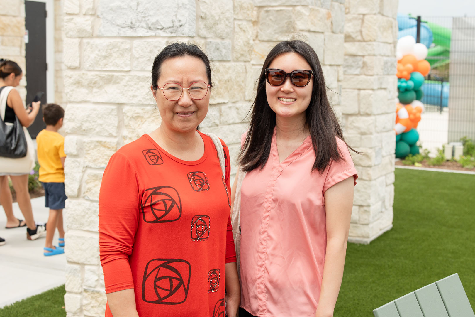 Two women stand side by side outdoors at the Haven Amenity Village Grand Opening, smiling at the camera. One wears a red dress and glasses, the other sports sunglasses and a pink blouse. Stone wall and balloons can be seen in the background.
