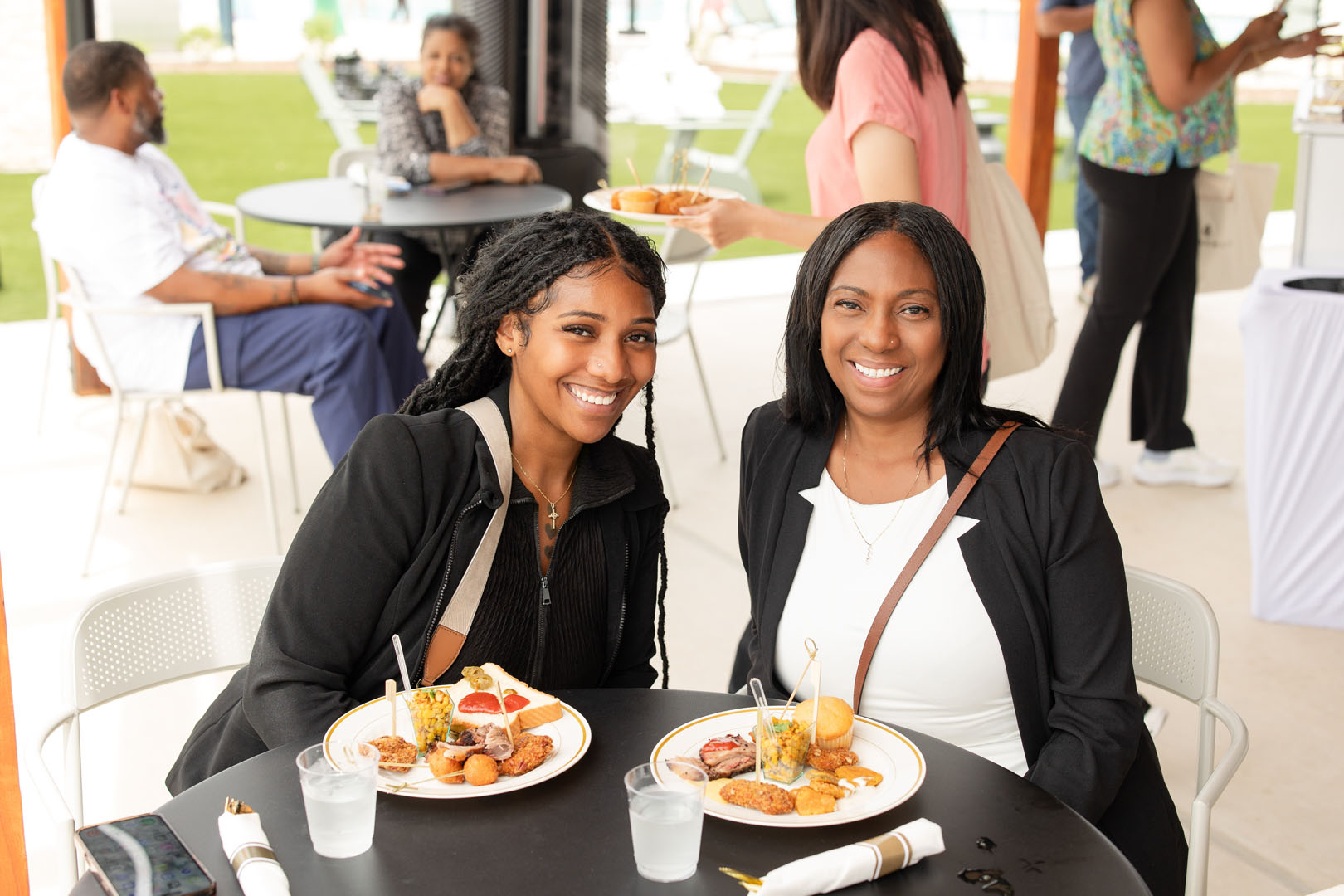 Two women sit at a table outdoors, smiling at the camera with plates of food and drinks in front of them during the Haven Amenity Village Grand Opening. Other people are seated or standing in the background.