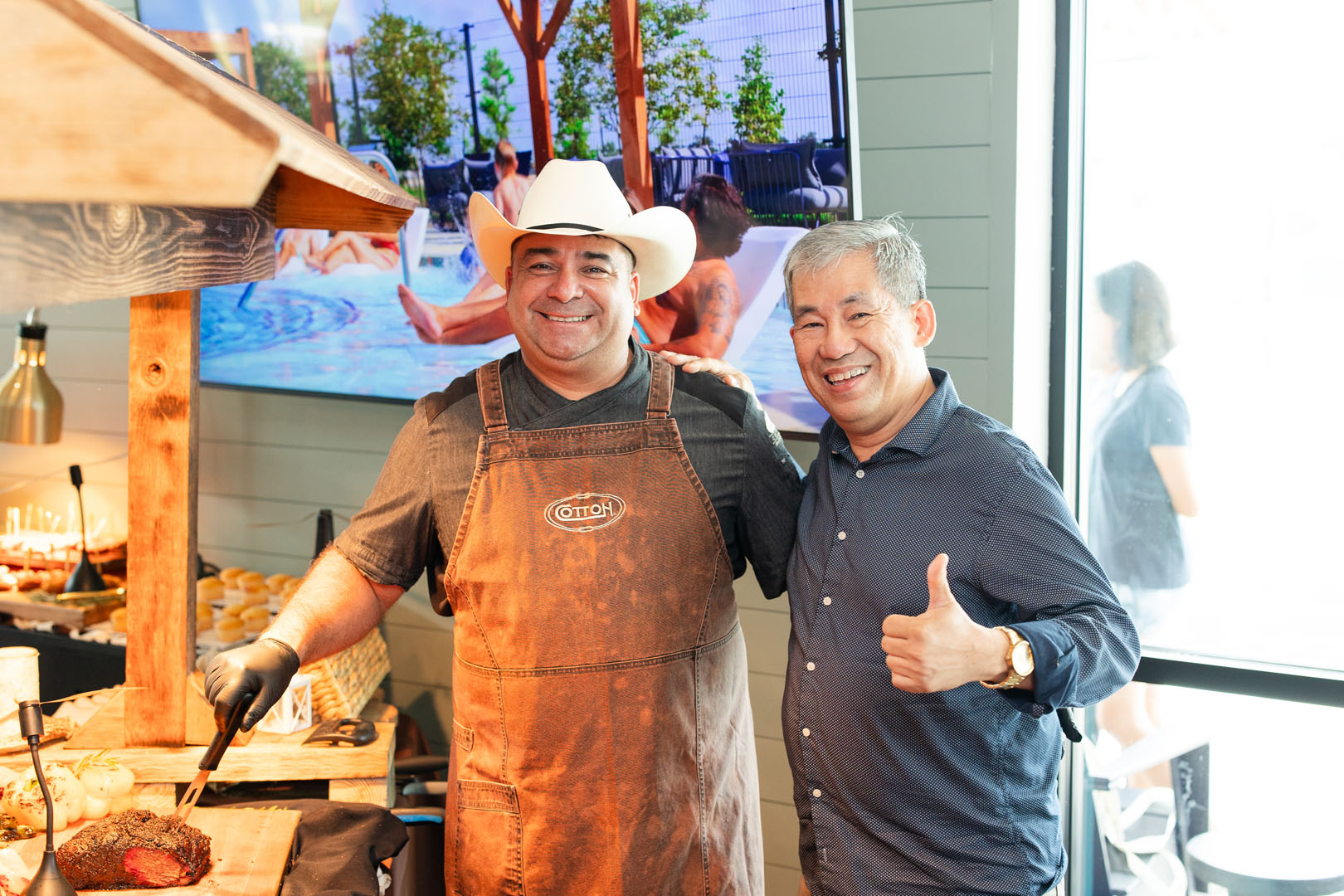 Two men stand indoors at Haven Amenity Village’s Grand Opening; one wears a cowboy hat and apron near food, while the other in a button-down shirt gives a thumbs-up. A large TV screen and window are visible in the background.