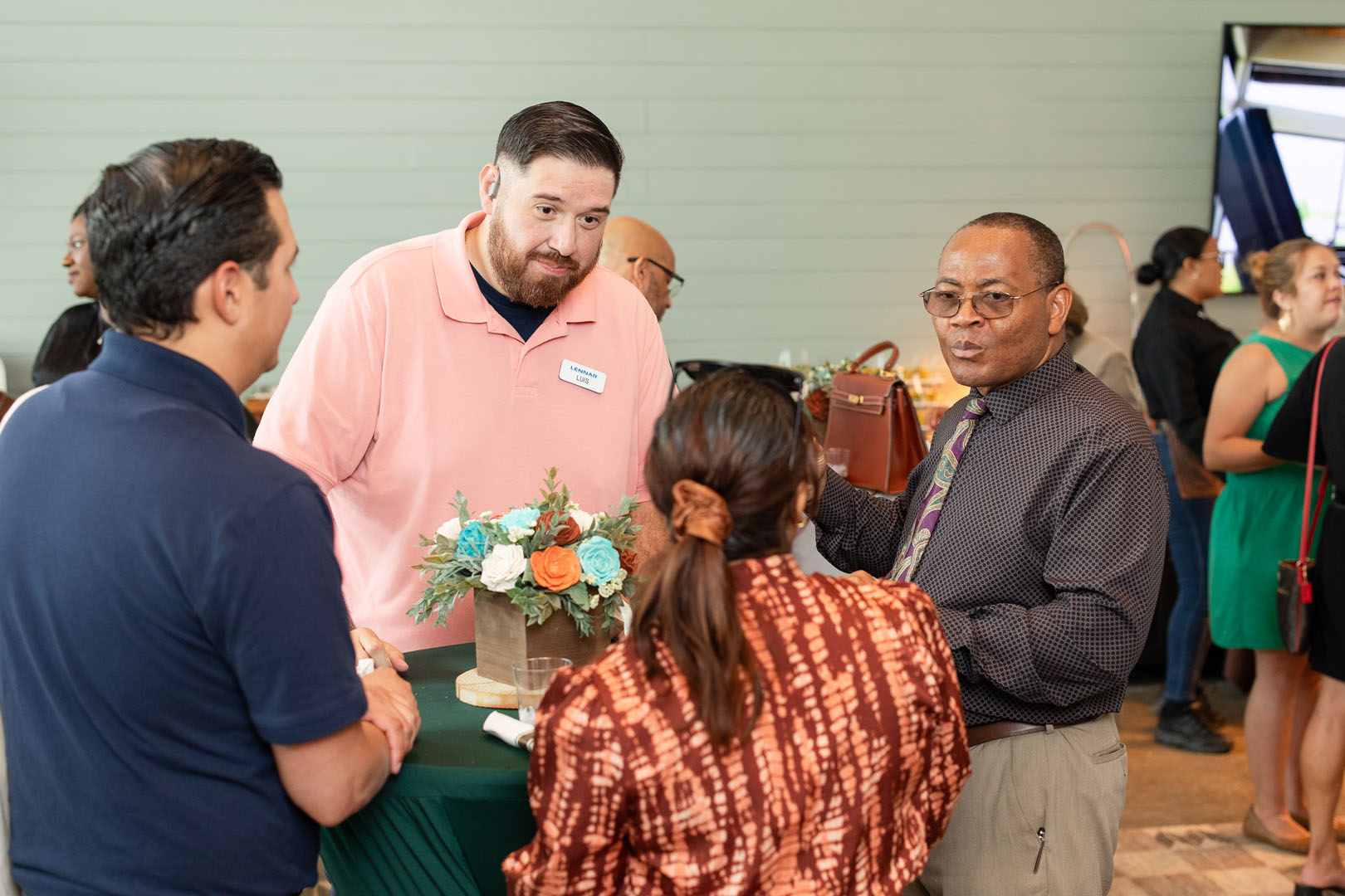 Four adults stand around a small table with a flower arrangement, engaged in conversation at the Grand Opening of Haven Amenity Village.