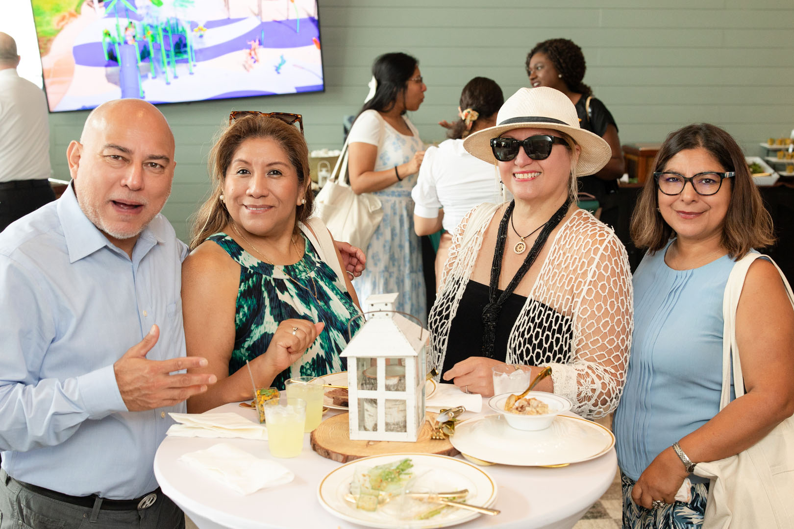 Four adults stand around a table with food and drinks, smiling at the camera during the Grand Opening of Haven Amenity Village. Other people are visible in the background, enjoying the indoor social event.