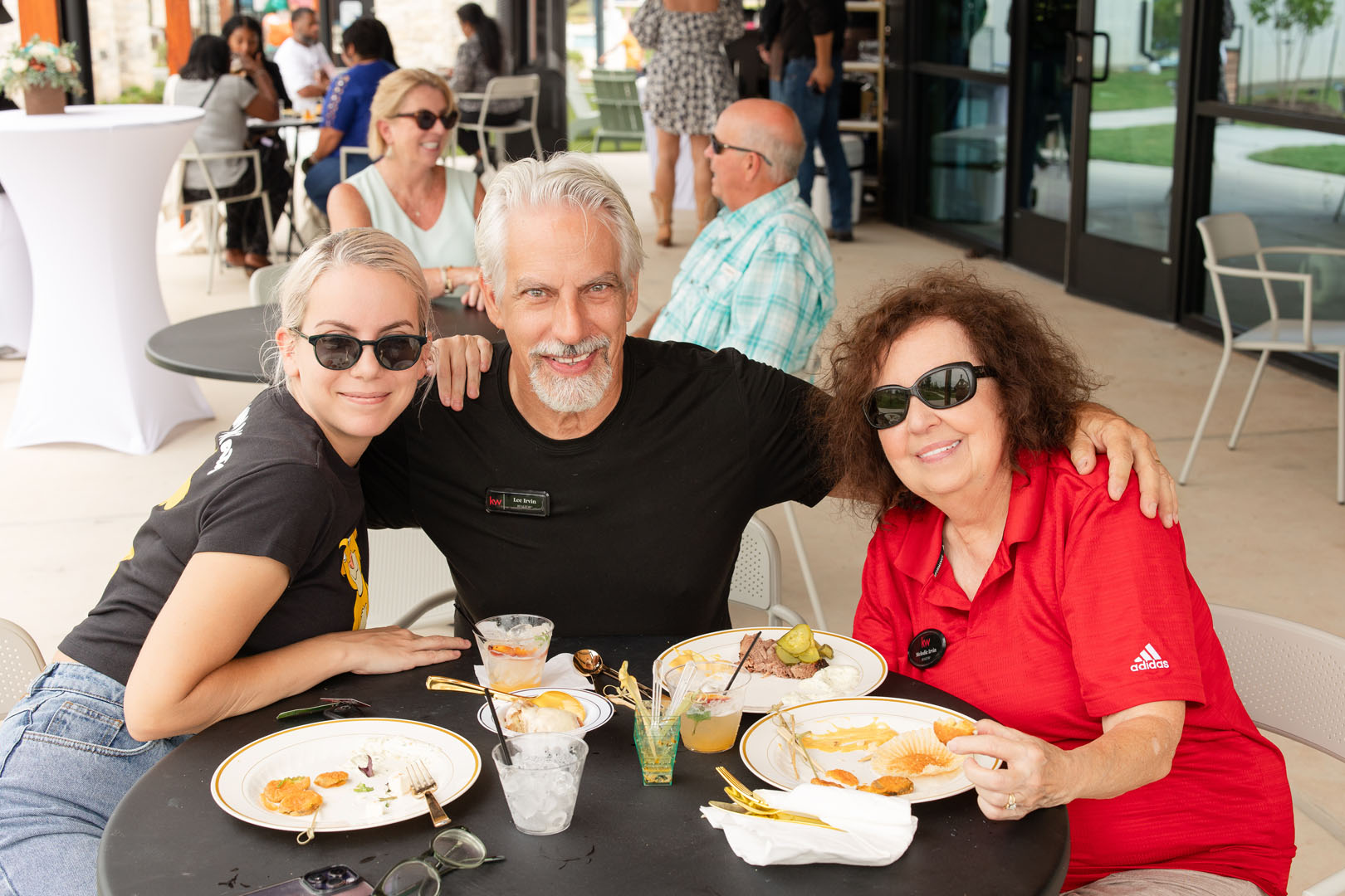Three adults wearing sunglasses sit at an outdoor table at Haven Amenity Village’s Grand Opening, smiling at the camera with plates of food. Other people are seated at tables in the background, enjoying the festive atmosphere.
