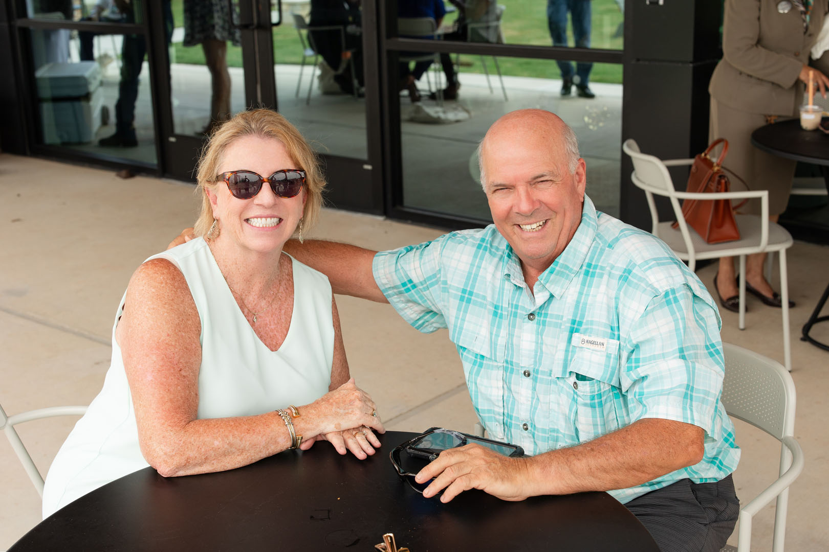 A woman and a man sit at an outdoor table, smiling at the camera during the Grand Opening of Haven Amenity Village. The man has his arm around the woman. Both are casually dressed and wearing sunglasses.