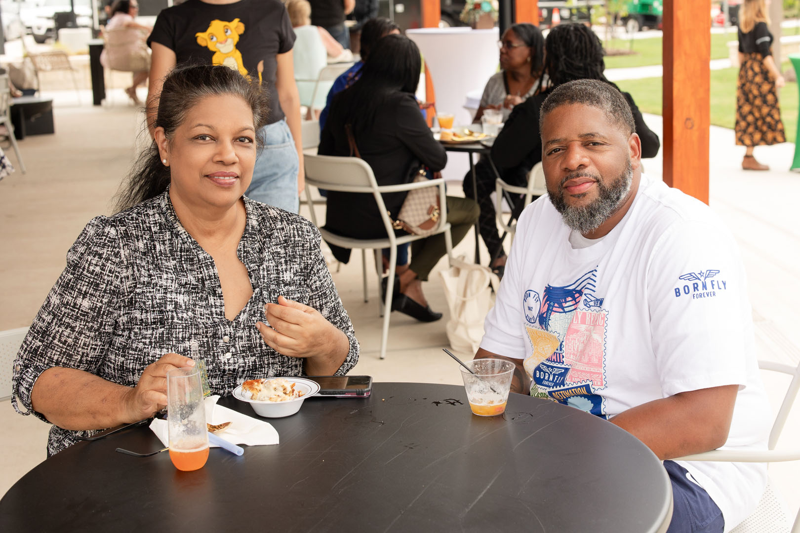 Two people sit at an outdoor table with drinks and food, looking toward the camera during the Grand Opening of Haven Amenity Village. Other guests can be seen enjoying the lively atmosphere in the background.