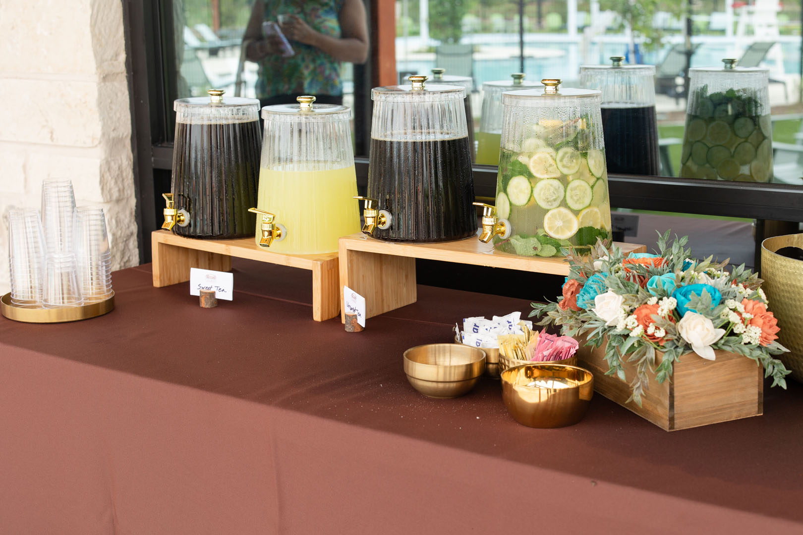 At the Haven Amenity Village Grand Opening, three beverage dispensers with iced tea, lemonade, and cucumber water sit on a brown tablecloth beside plastic cups, gold bowls, and a box of colorful artificial flowers.
