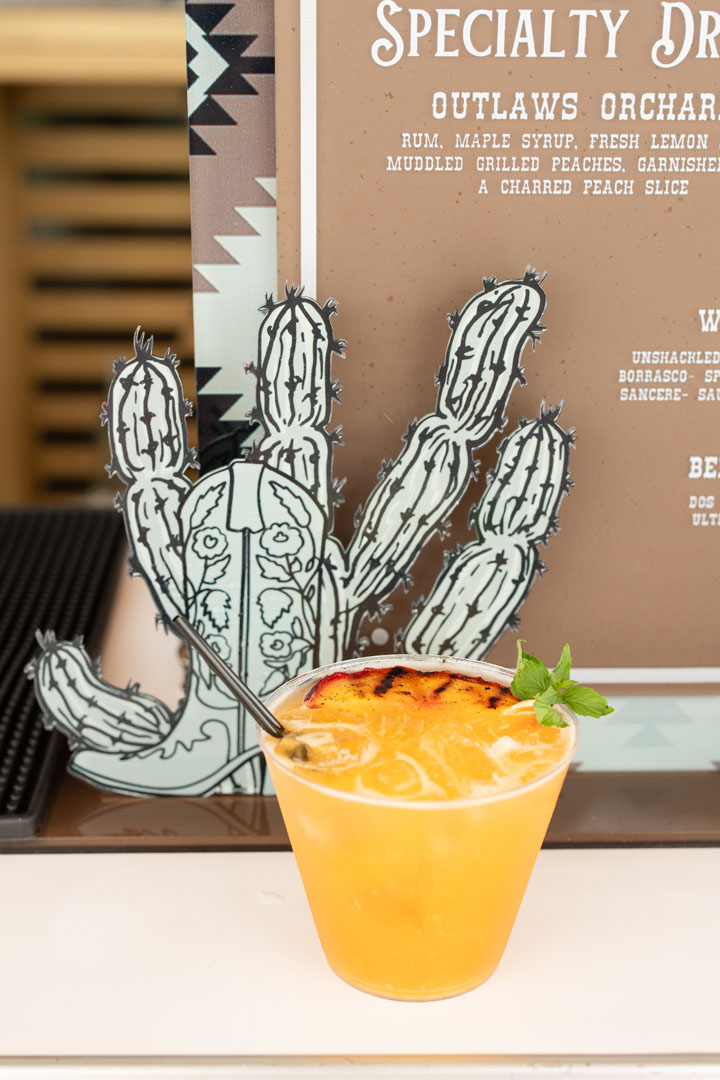 A yellow-orange cocktail garnished with a charred peach slice and mint sits in front of a menu with a cactus illustration, celebrating the Grand Opening of Haven Amenity Village.