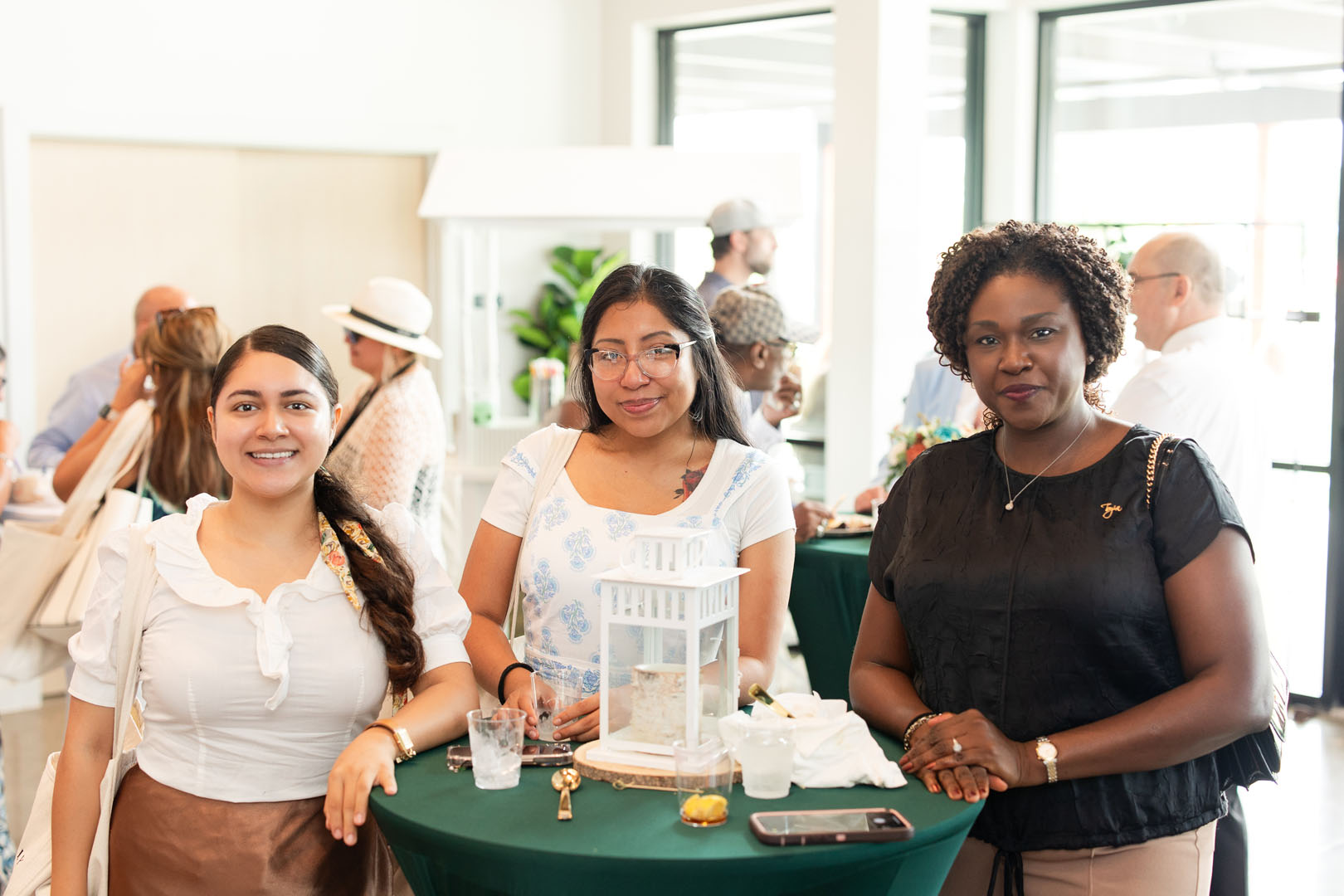 Three women stand around a green cocktail table at the Haven Amenity Village Grand Opening, smiling at the camera during an indoor social event, with other guests mingling in the background.