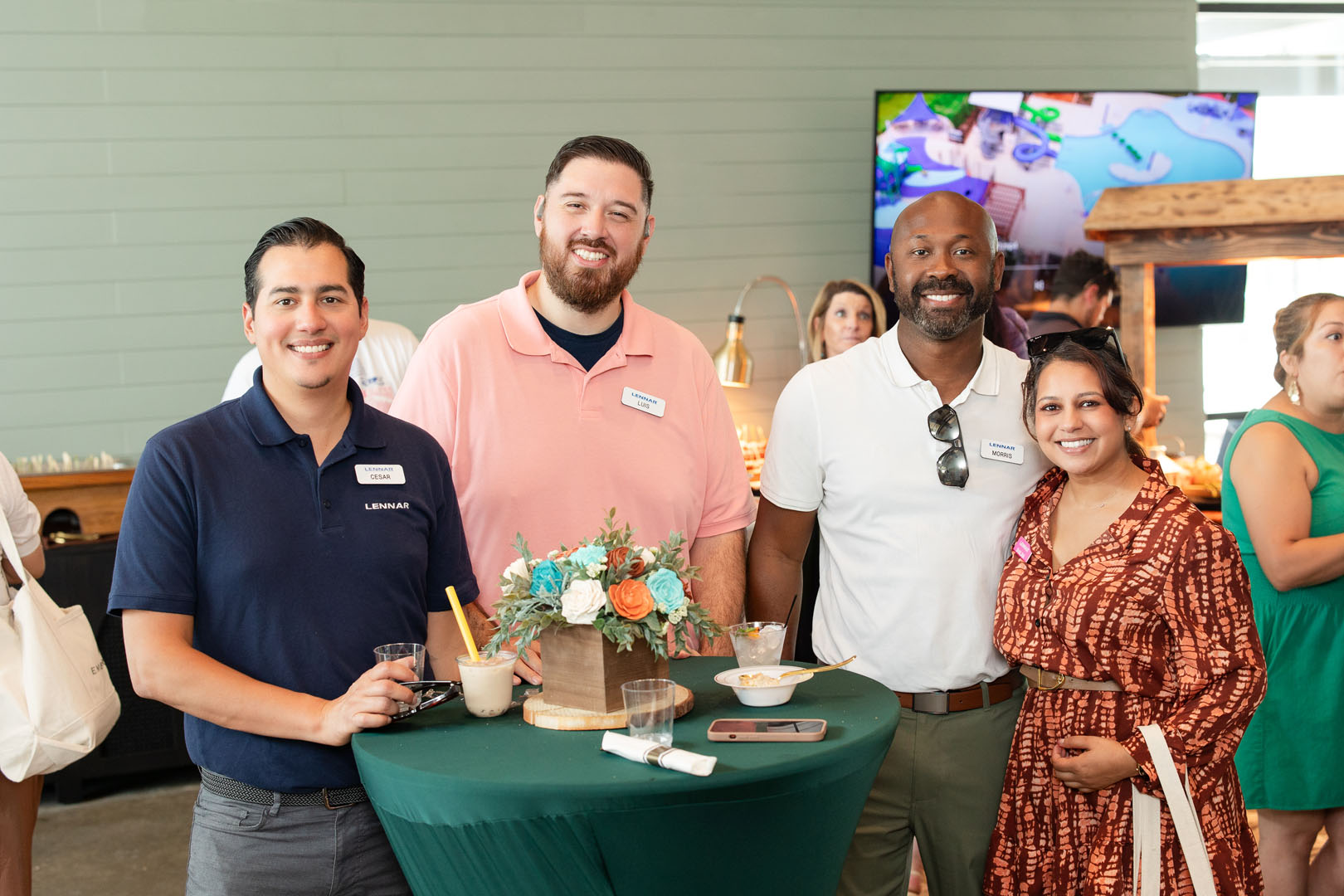 Four people stand around a green cocktail table with drinks and snacks at the Haven Amenity Village Grand Opening, smiling at the camera during this lively indoor social event.