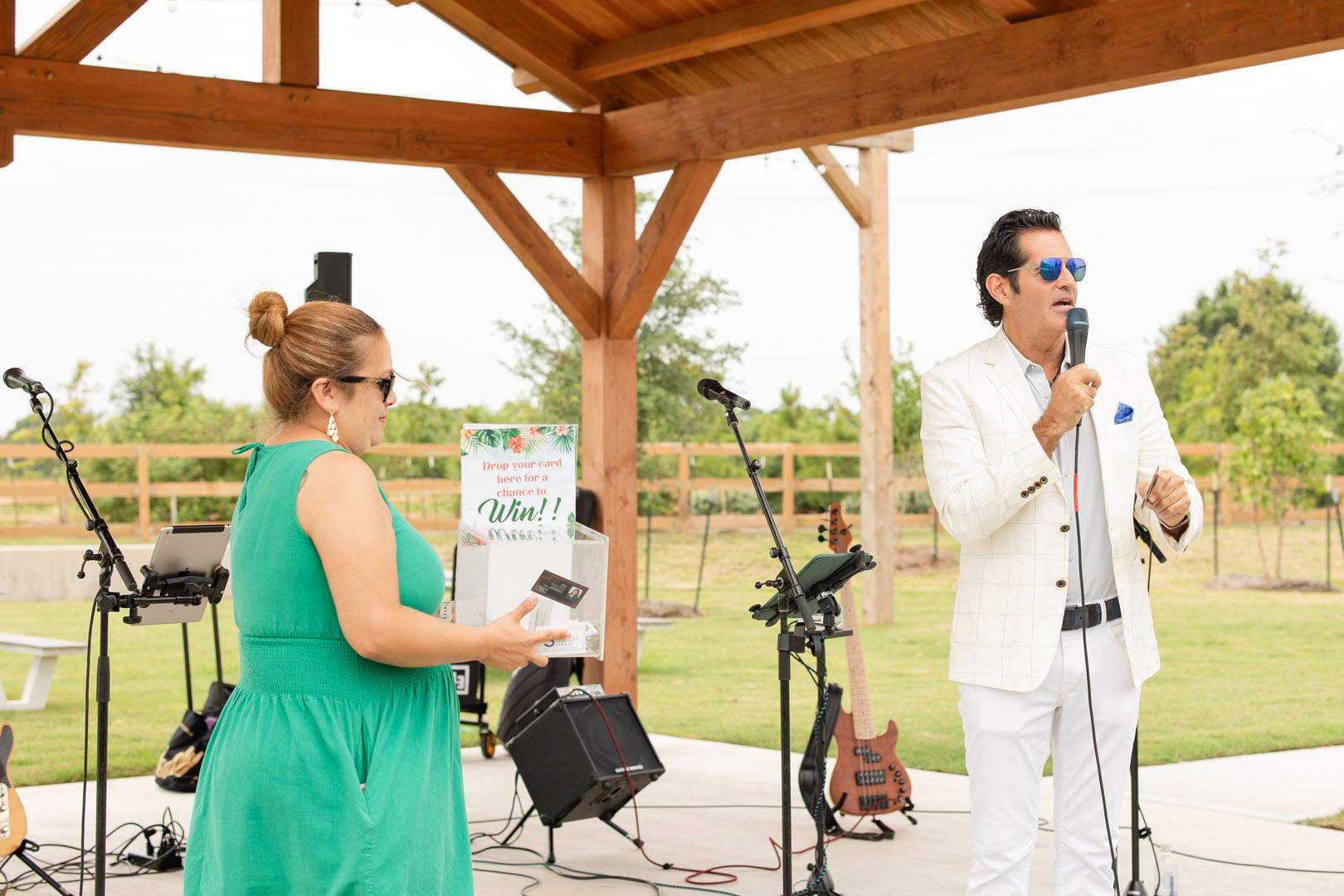 At the Haven Amenity Village Grand Opening, a man in a white suit speaks into a microphone while a woman in a green dress holds a box at an outdoor event under a wooden pavilion.