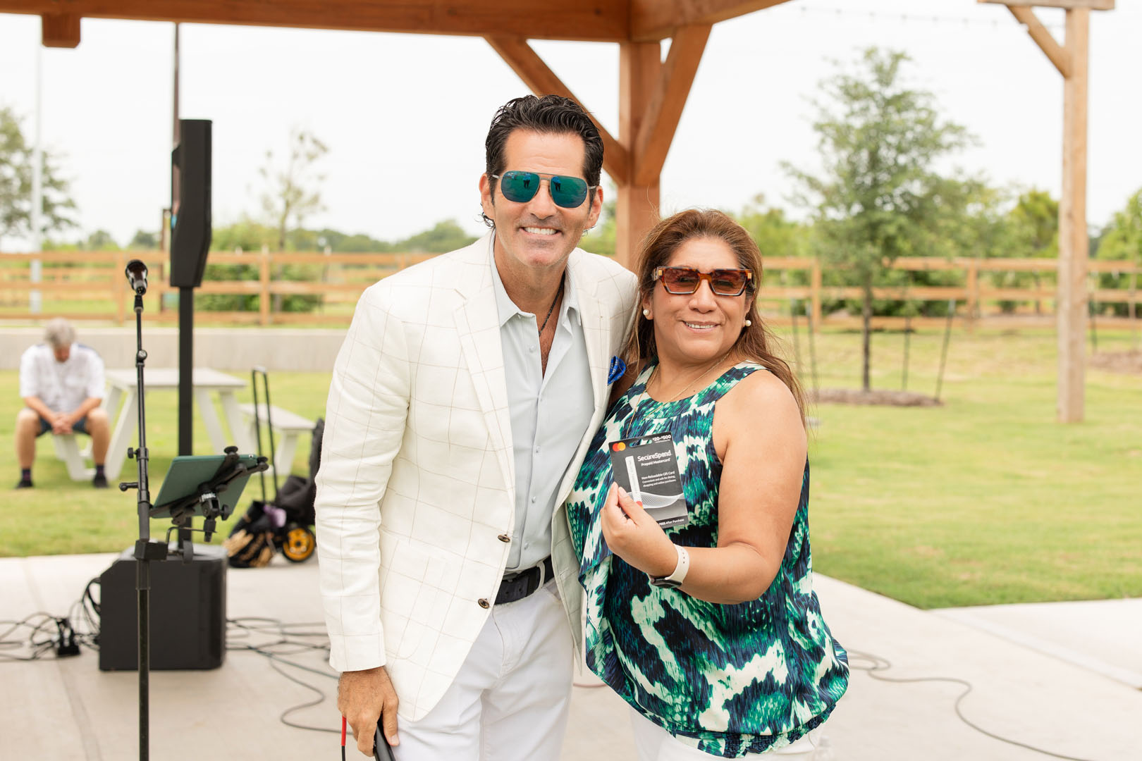 A man in a white suit and a woman in a blue patterned top stand side by side outdoors at the Haven Amenity Village Grand Opening, both smiling and wearing sunglasses. The woman proudly holds a small plaque or award.