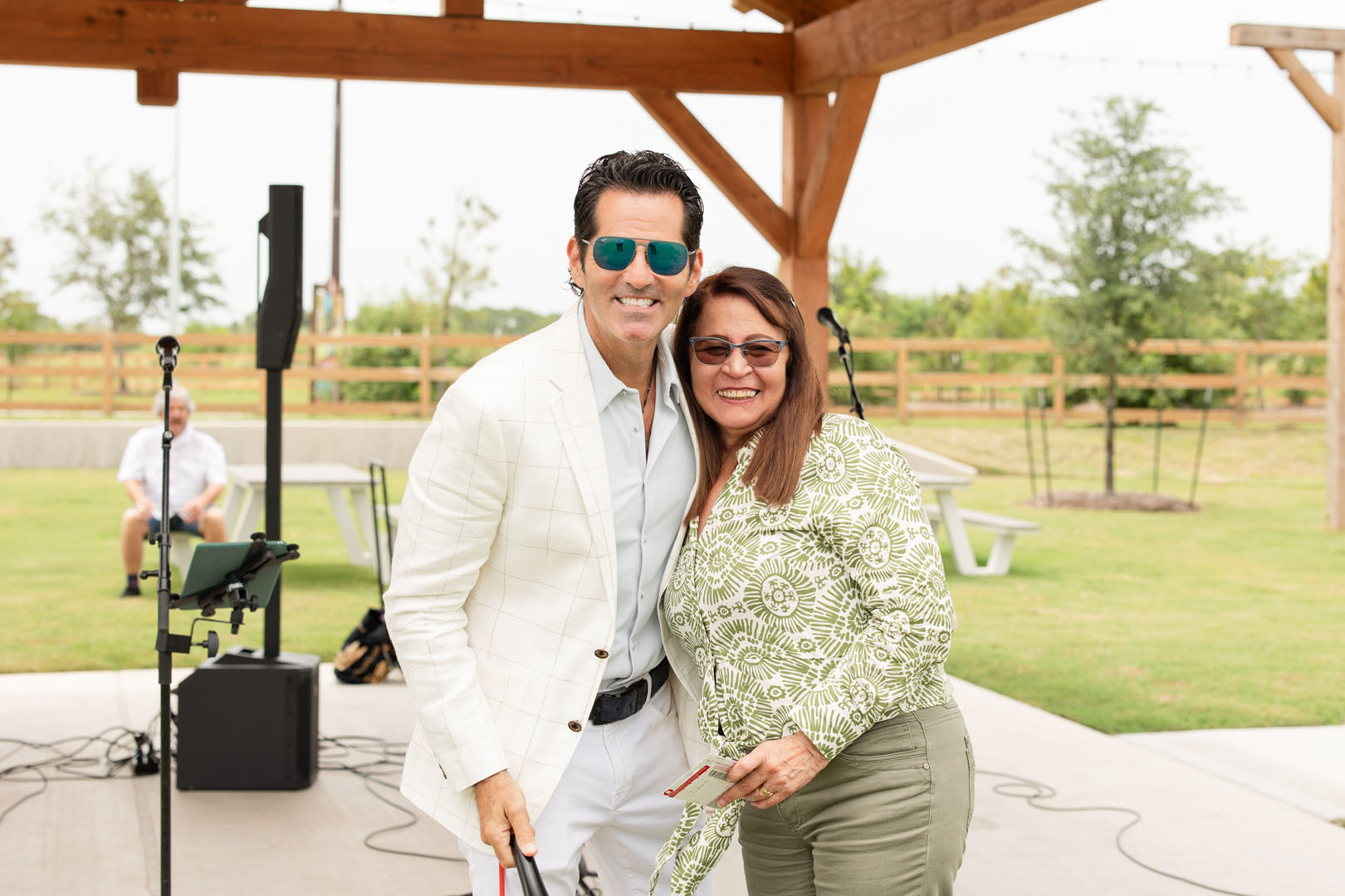 A man and a woman pose and smile under a wooden pavilion at the Haven Amenity Village Grand Opening, with grass, picnic tables, and audio equipment visible in the background.
