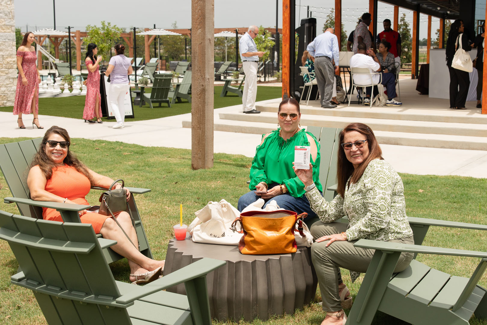 Three women sit and smile around a table outdoors at the Grand Opening of Haven Amenity Village, with drinks and bags, while others mingle in the background near a covered patio area.