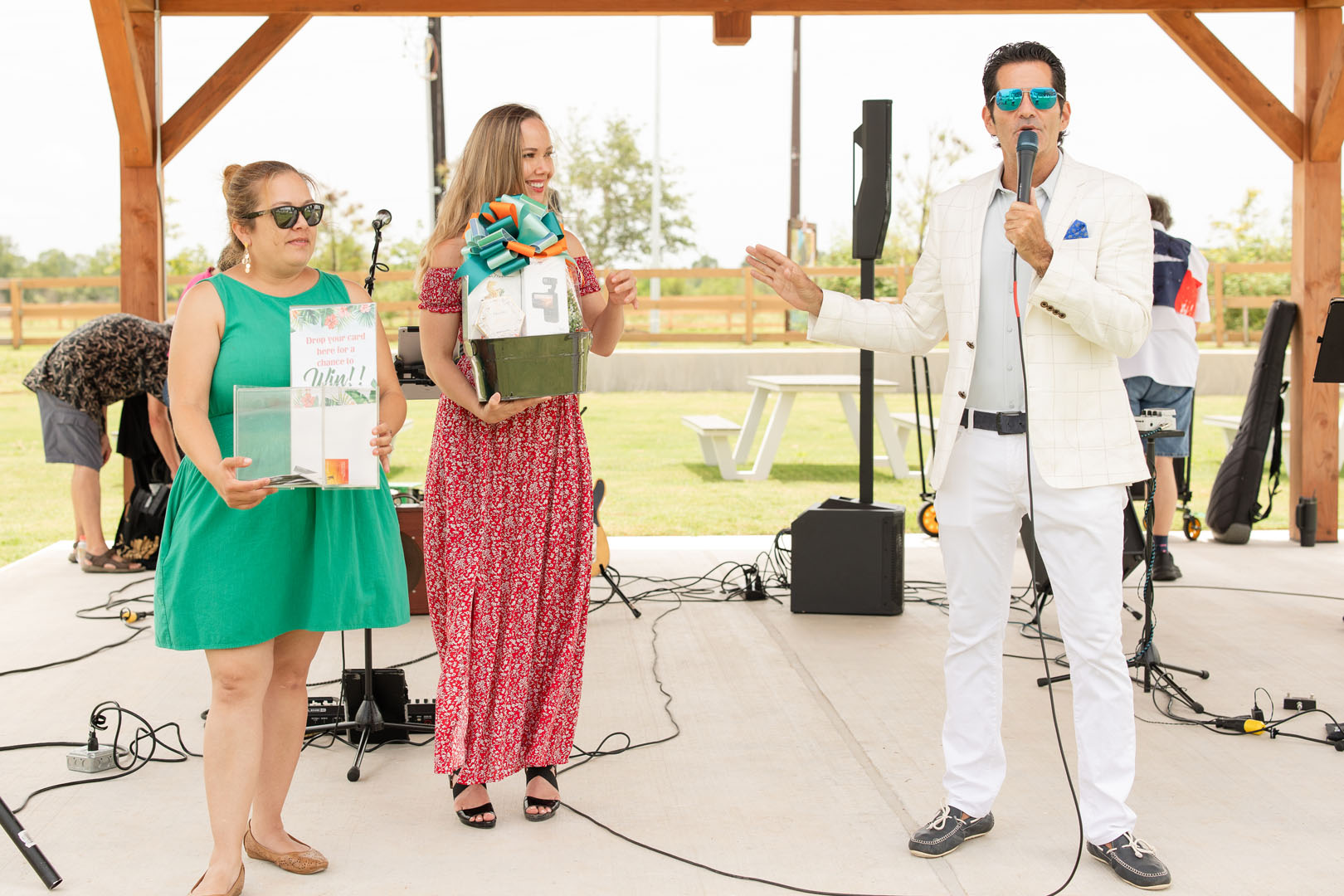 At the Haven Amenity Village Grand Opening, a man in a white suit speaks into a microphone while two women stand beside him—one holding a gift basket and the other with a clear gift bag—on an outdoor stage.