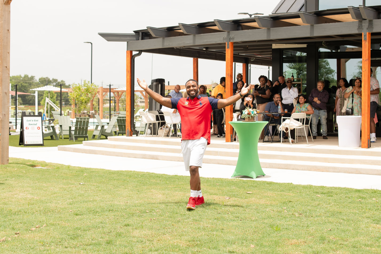 A man in athletic wear smiles with arms raised outside a building at the Haven Amenity Village Grand Opening, while a group of people watches from a patio in the background.