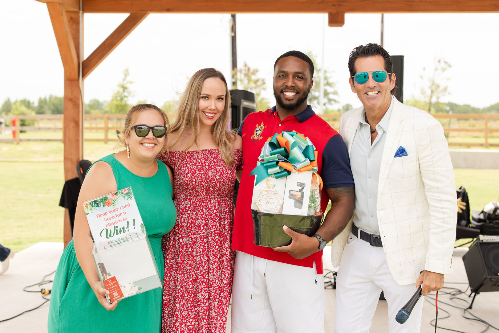 Four people stand together outdoors at the Grand Opening of Haven Amenity Village; one woman holds a sign, another stands beside her, a man holds a gift basket, and another man in white holds a microphone.