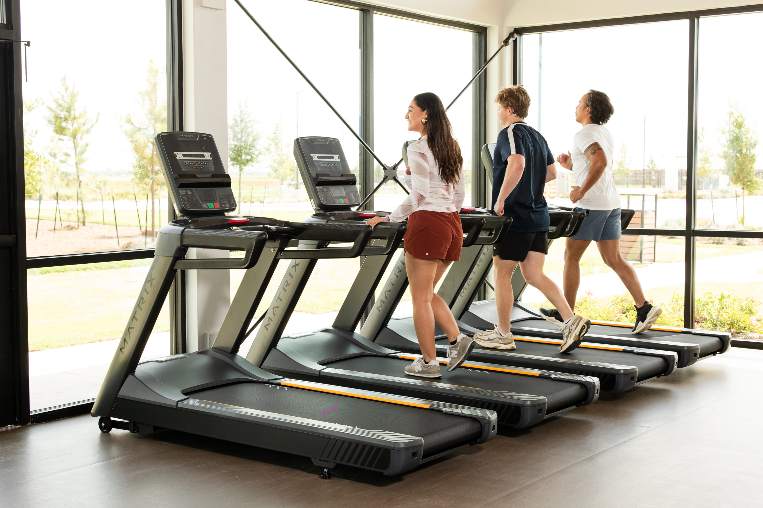 Three people are exercising on treadmills in a bright gym with large windows at The Haven Amenity Village, each facing forward as they walk or run.