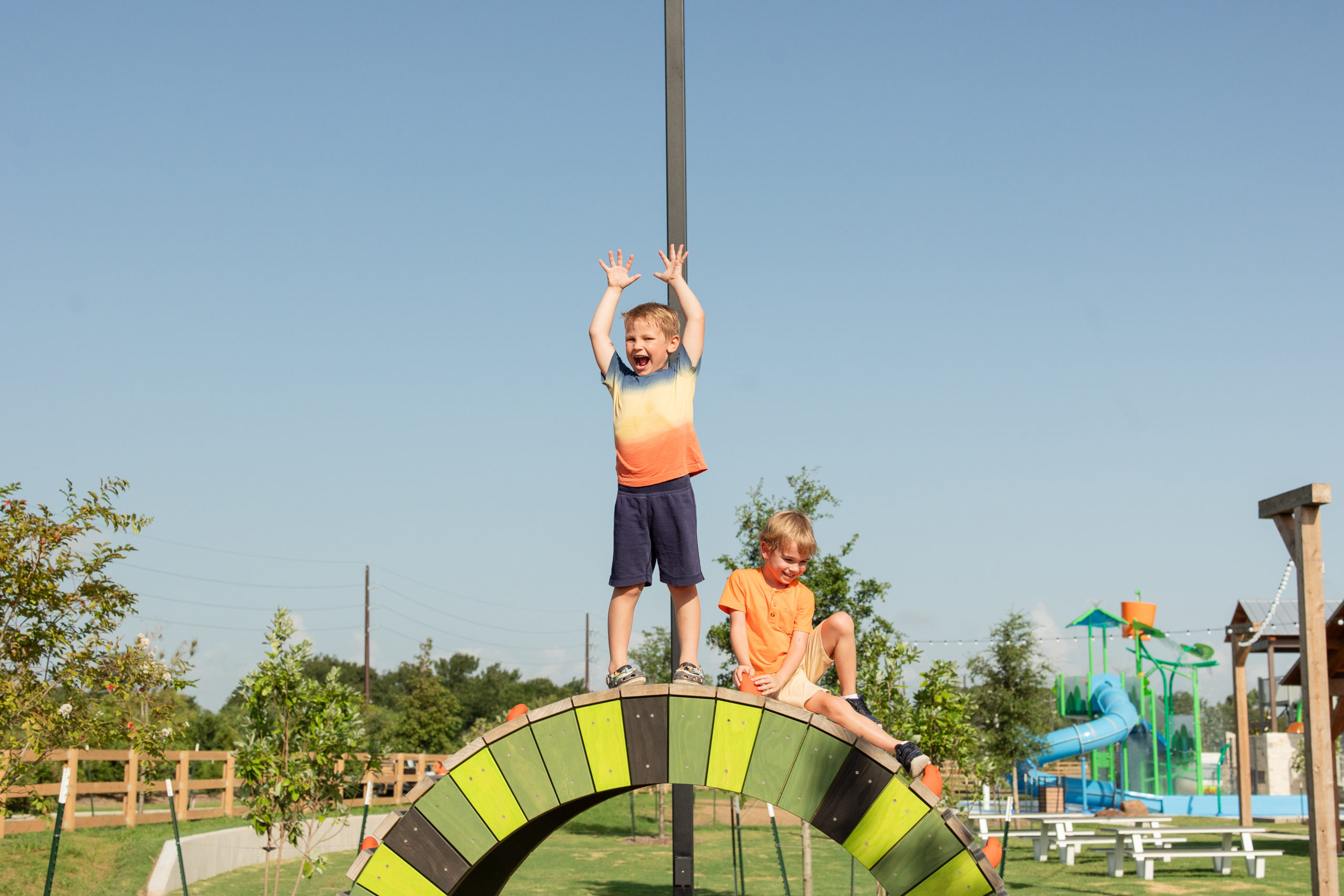 Two children play on top of a colorful arch structure at The Haven Amenity Village; one stands with arms raised while the other sits, with playground equipment and greenery in the background.