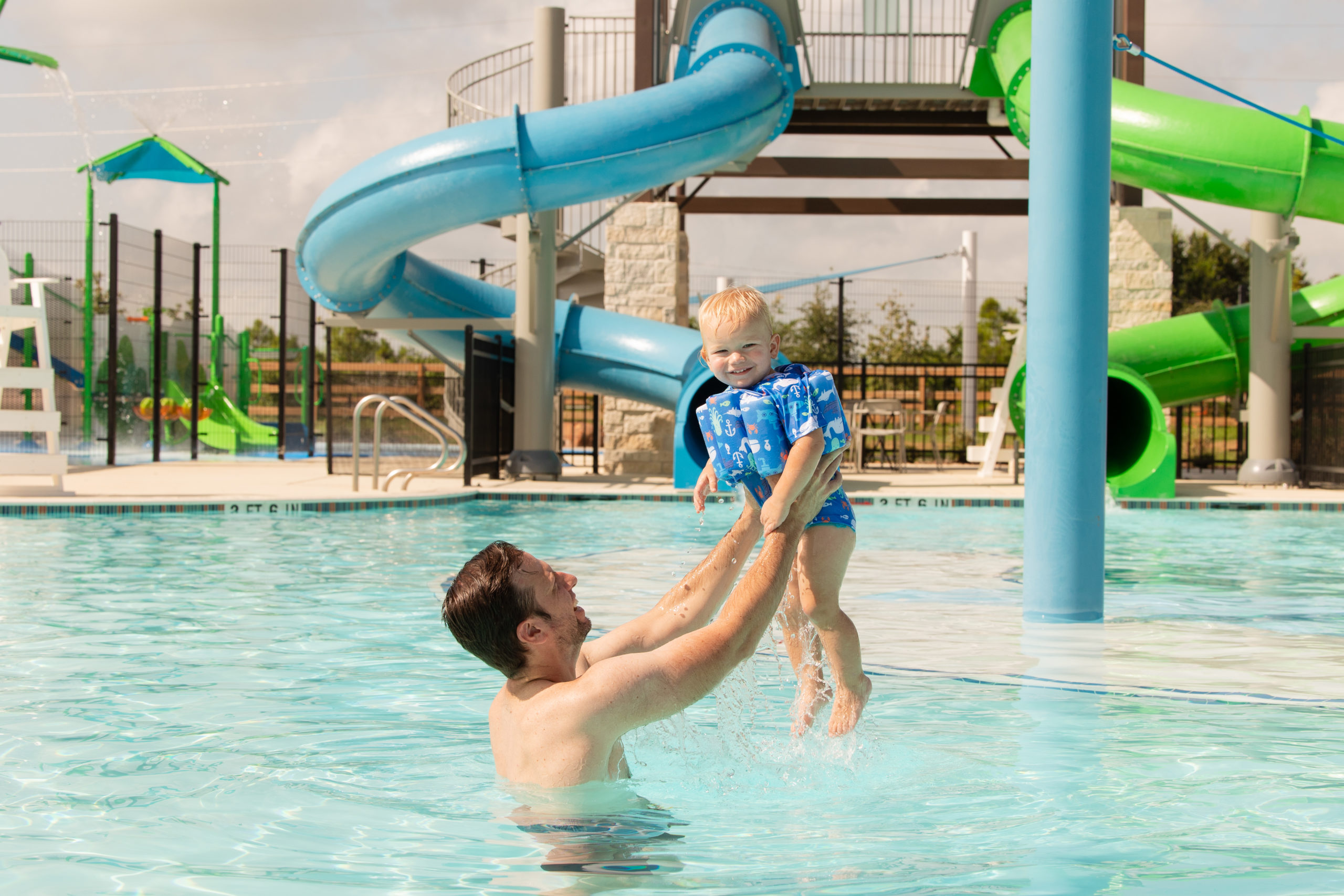 An adult lifts a smiling toddler wearing a blue flotation vest in a swimming pool at Amenity Village, with water slides and endless fun in the background.